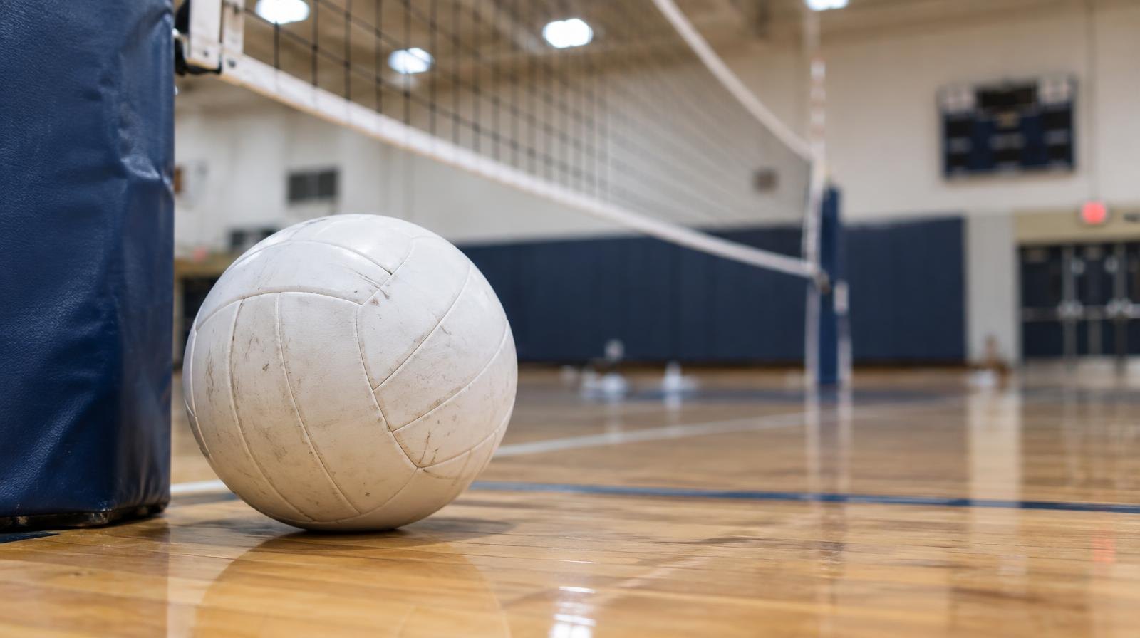 A white volleyball on a hardwood gym floor beside a volleyball net.