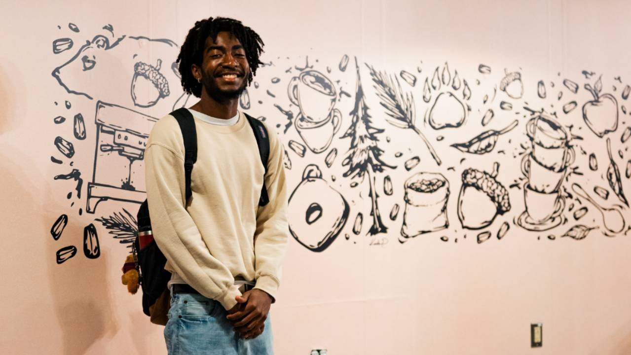 Smiling young man in a cream sweatshirt stands before a wall of hand-drawn woodland illustrations.