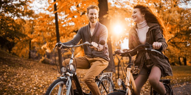 Two adults smile while riding bicycles side by side on a tree-lined path with orange and red autumn foliage.