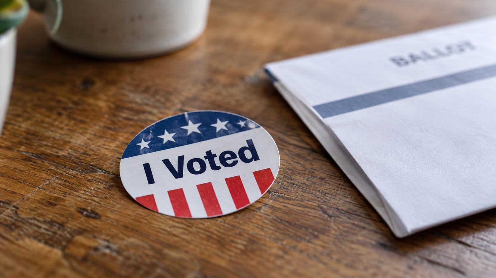An I Voted sticker resting on a wooden surface beside a folded ballot envelope.