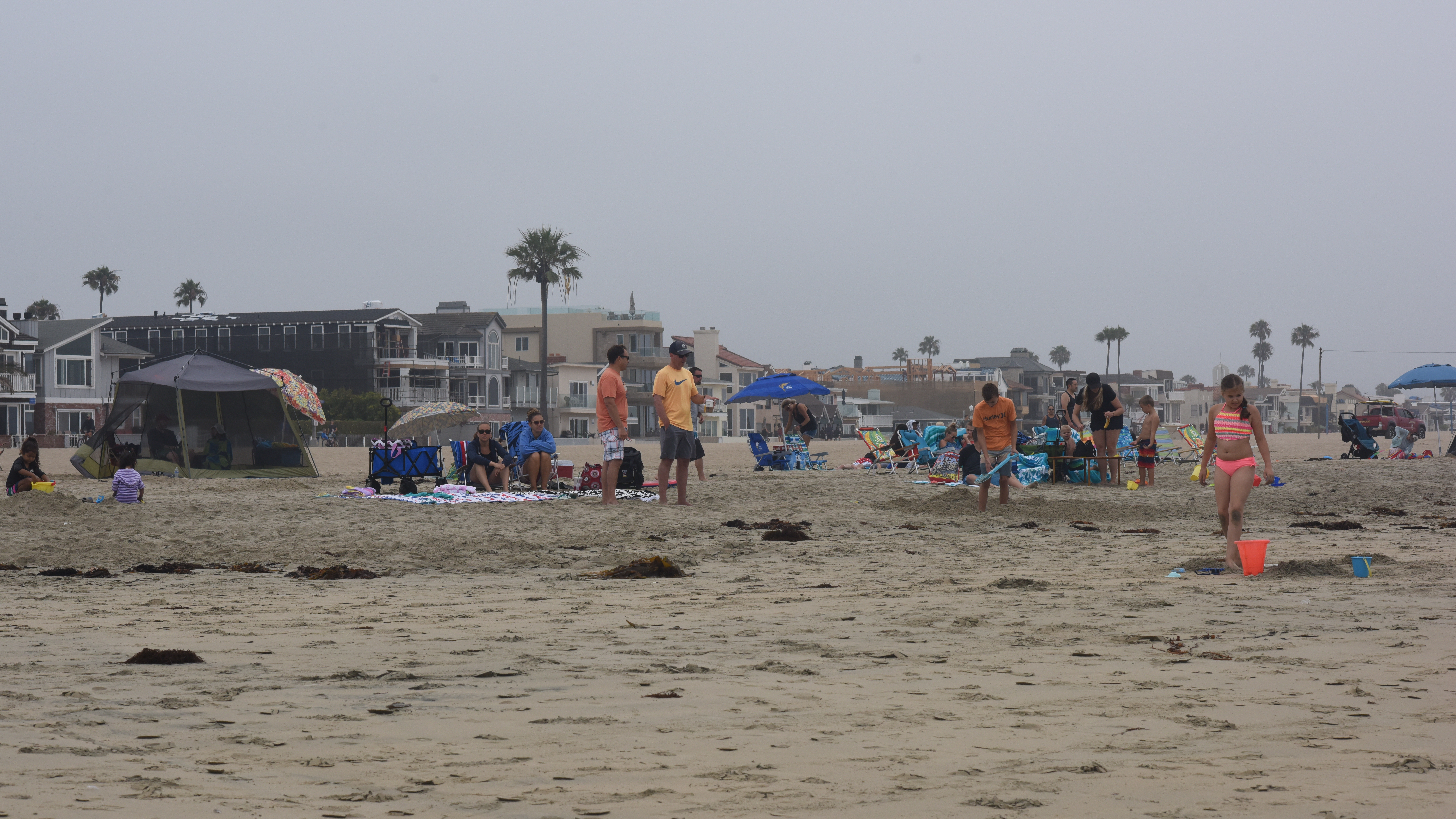 Crowded sandy beach with a large dark canopy tent and blue umbrellas set up near the water.