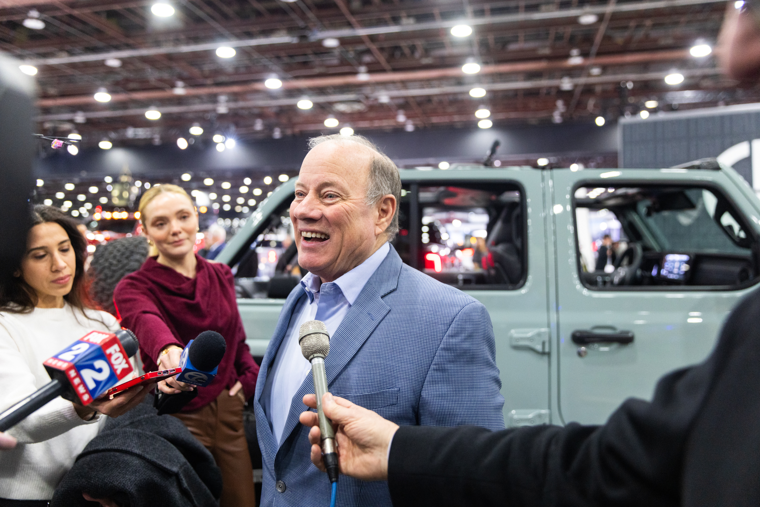 Man in blue blazer speaking into microphones at an indoor auto show event.