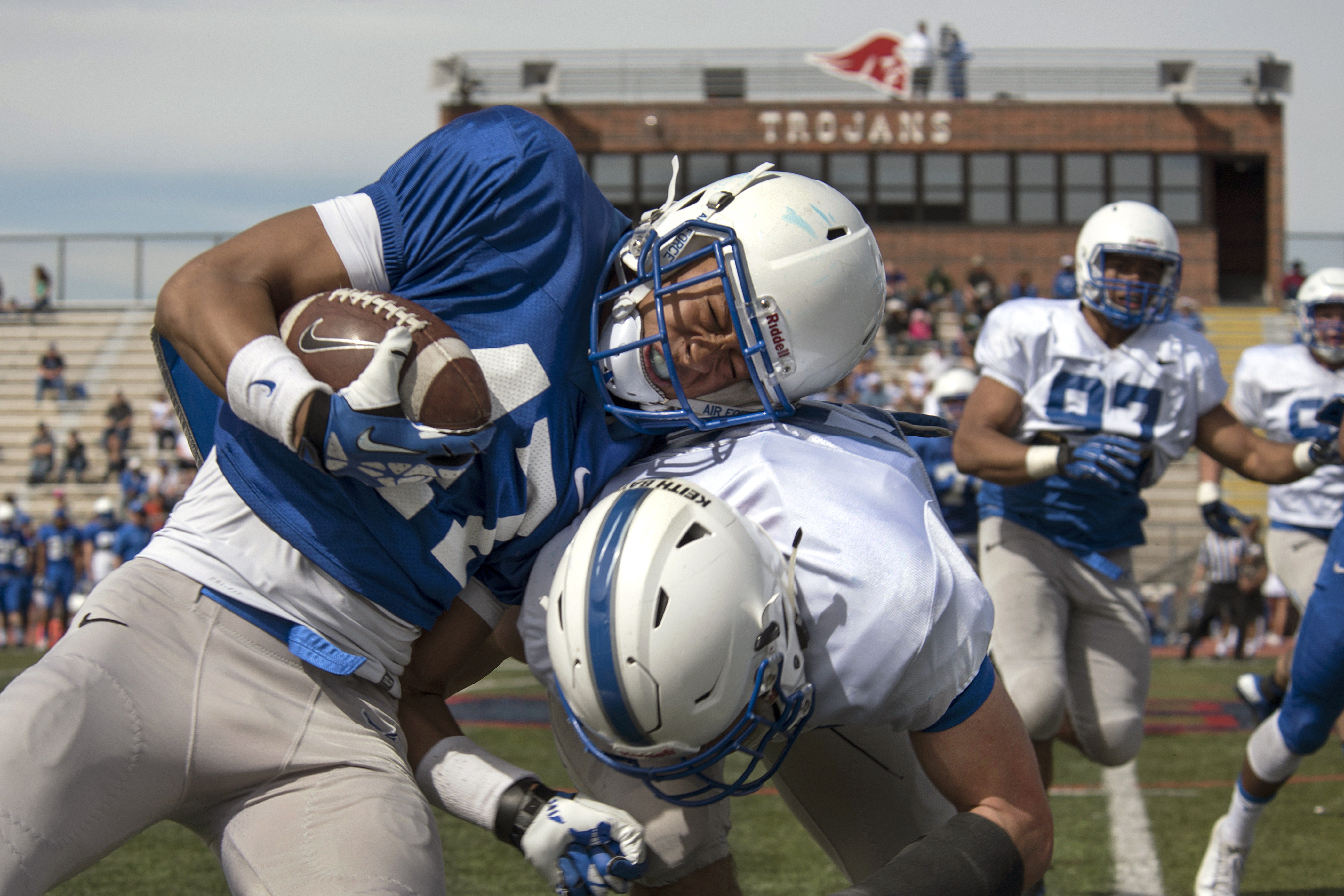 Football players in blue and white uniforms collide during a scrimmage on a grass field.