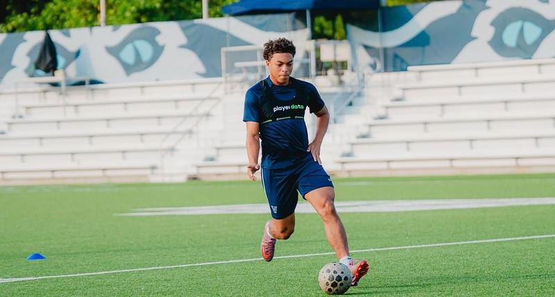 A soccer player in a navy kit dribbles a ball on an artificial turf field.