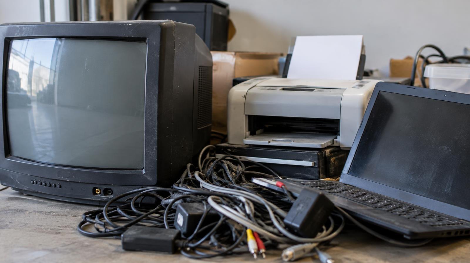 A pile of old electronics including a CRT television, laptop, printer, and cables stacked together.