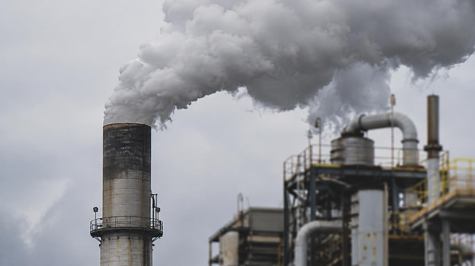 Industrial smokestack releasing thick smoke plumes against a hazy overcast sky.