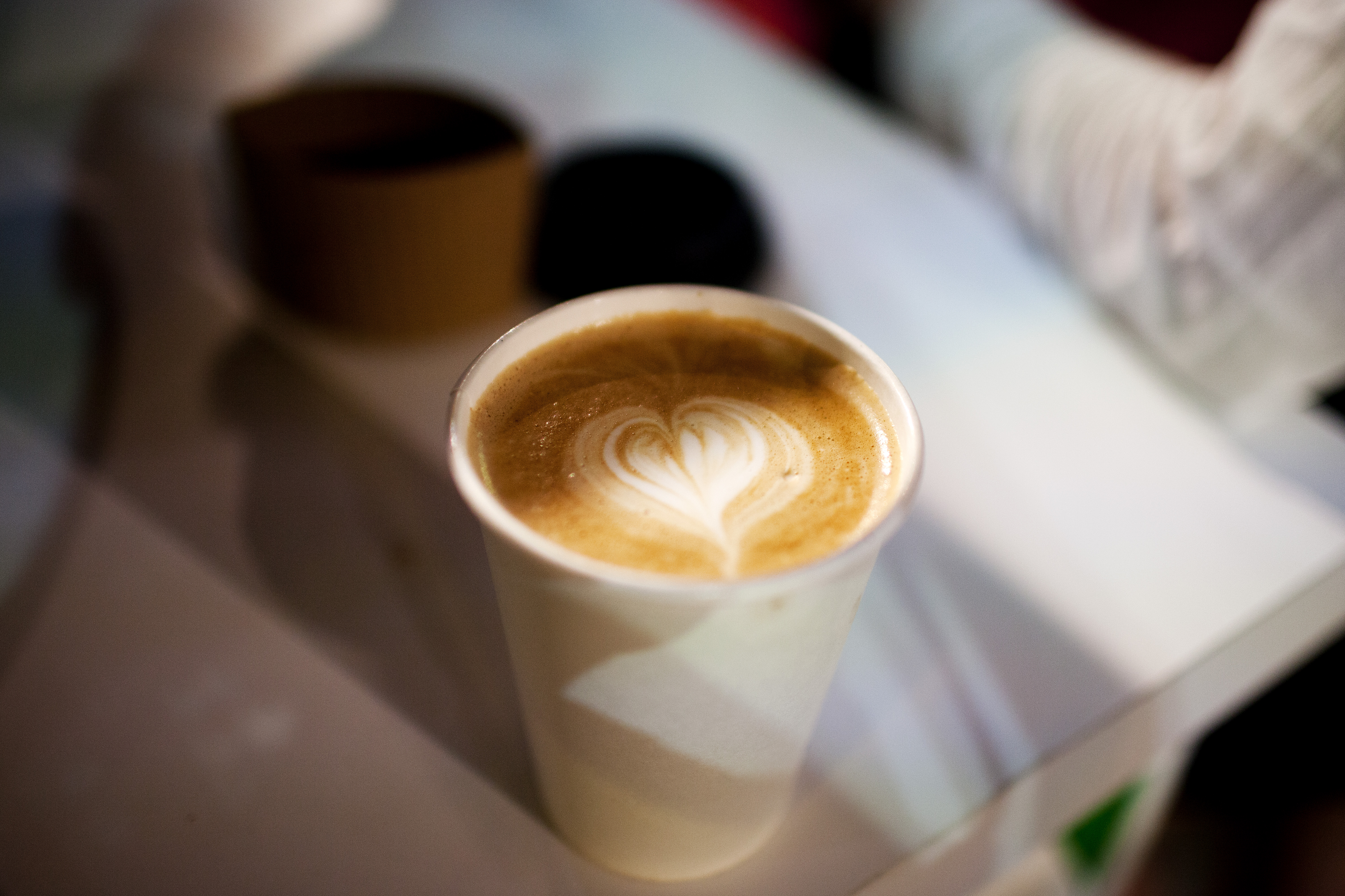 A paper cup of latte with heart-shaped latte art on a white surface.
