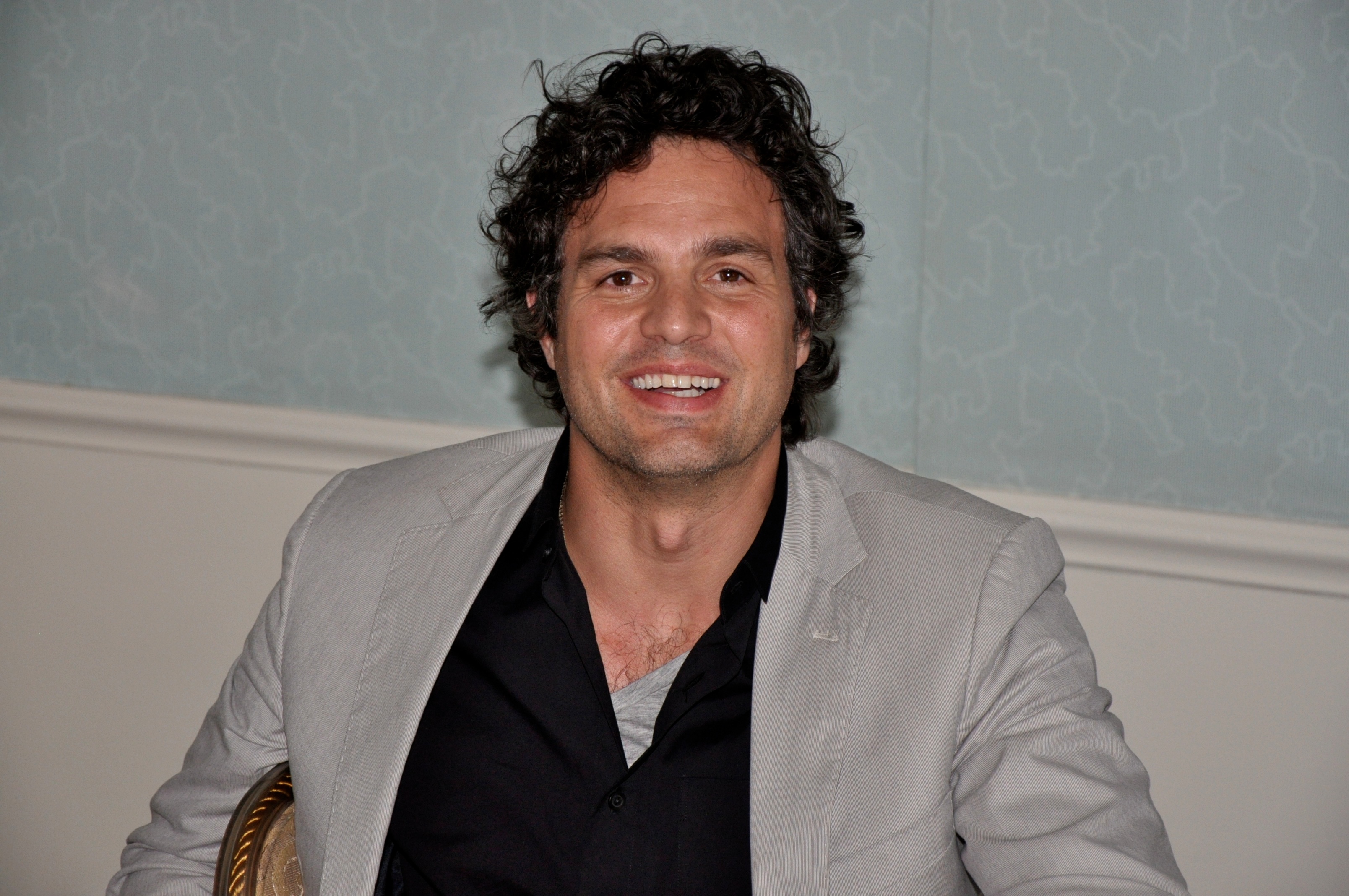 A smiling man with curly dark hair in a grey blazer seated indoors against a blue patterned wall.