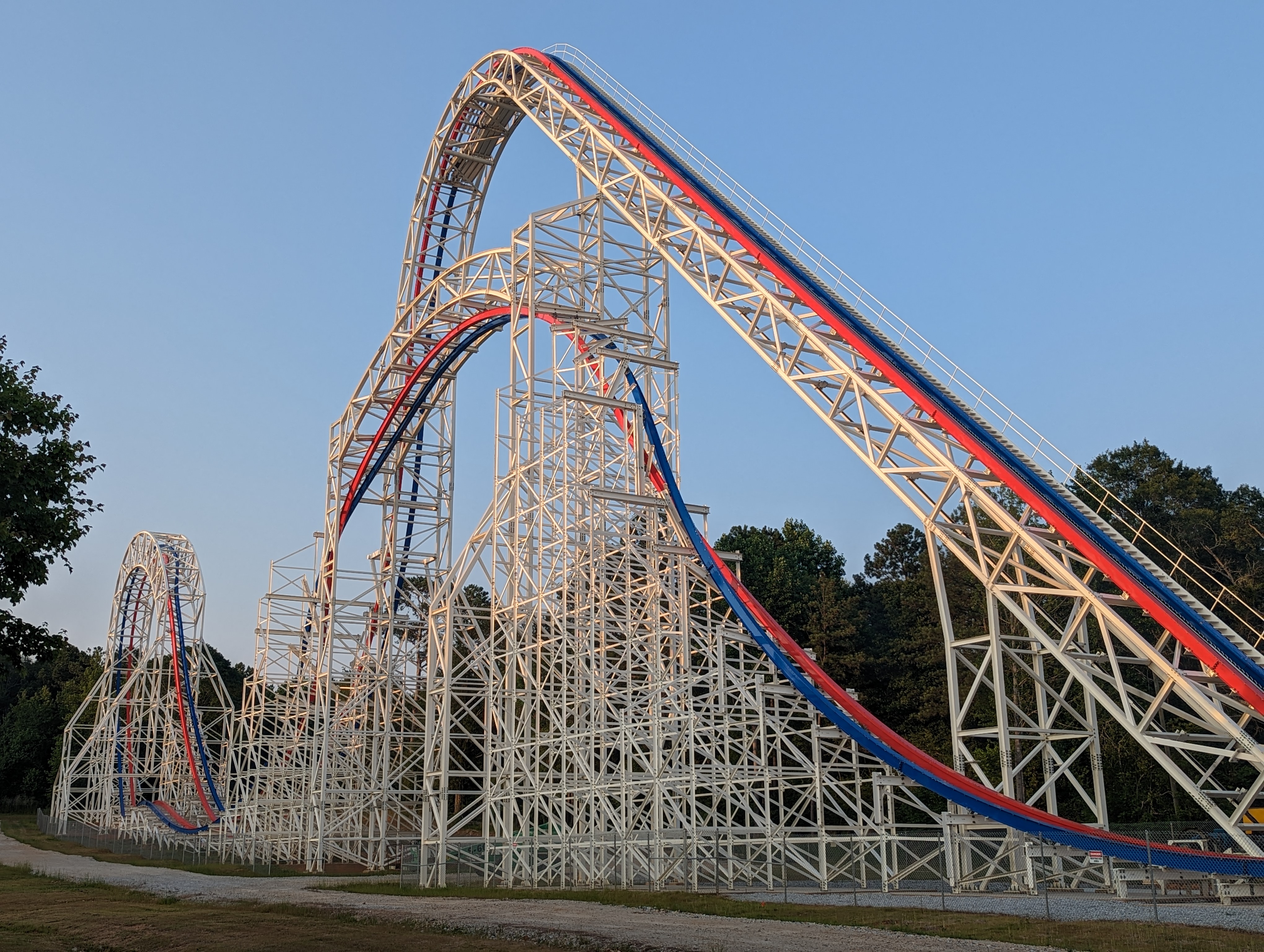 Hybrid roller coaster with red and blue steel track rising against a blue sky.