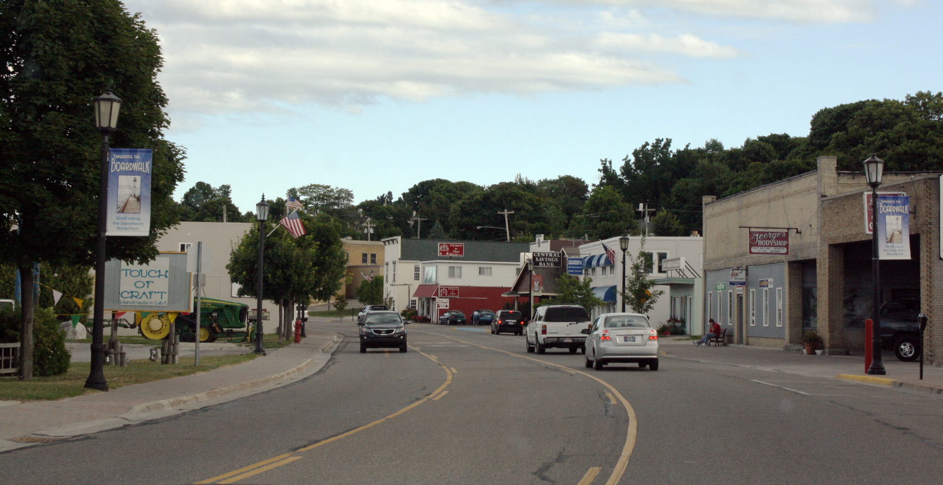 Main street of a small town with storefronts, banner-lined lamp posts, and passing cars.