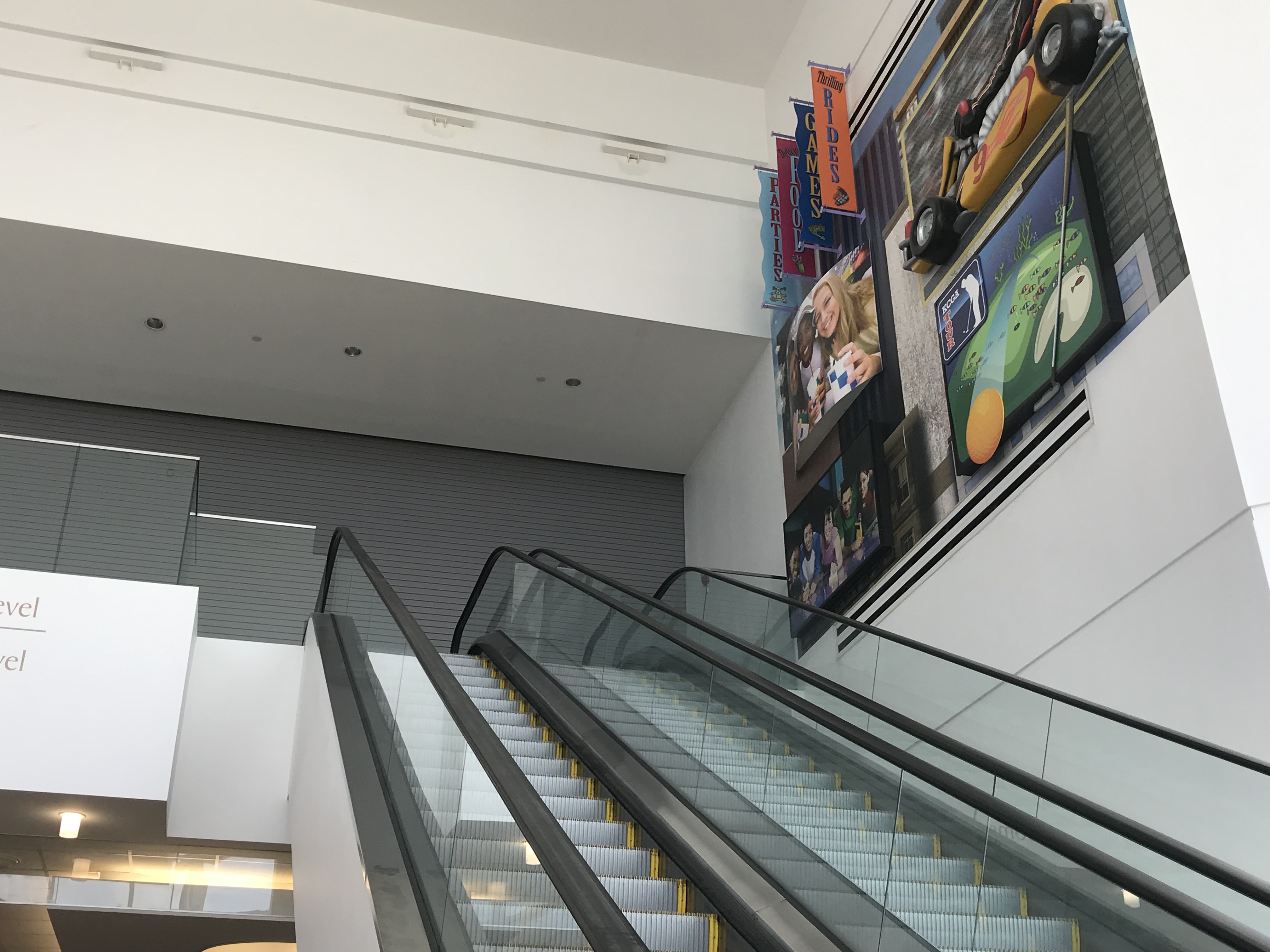 Mall interior with two escalators, colorful banners, and a large photo mural on the wall.