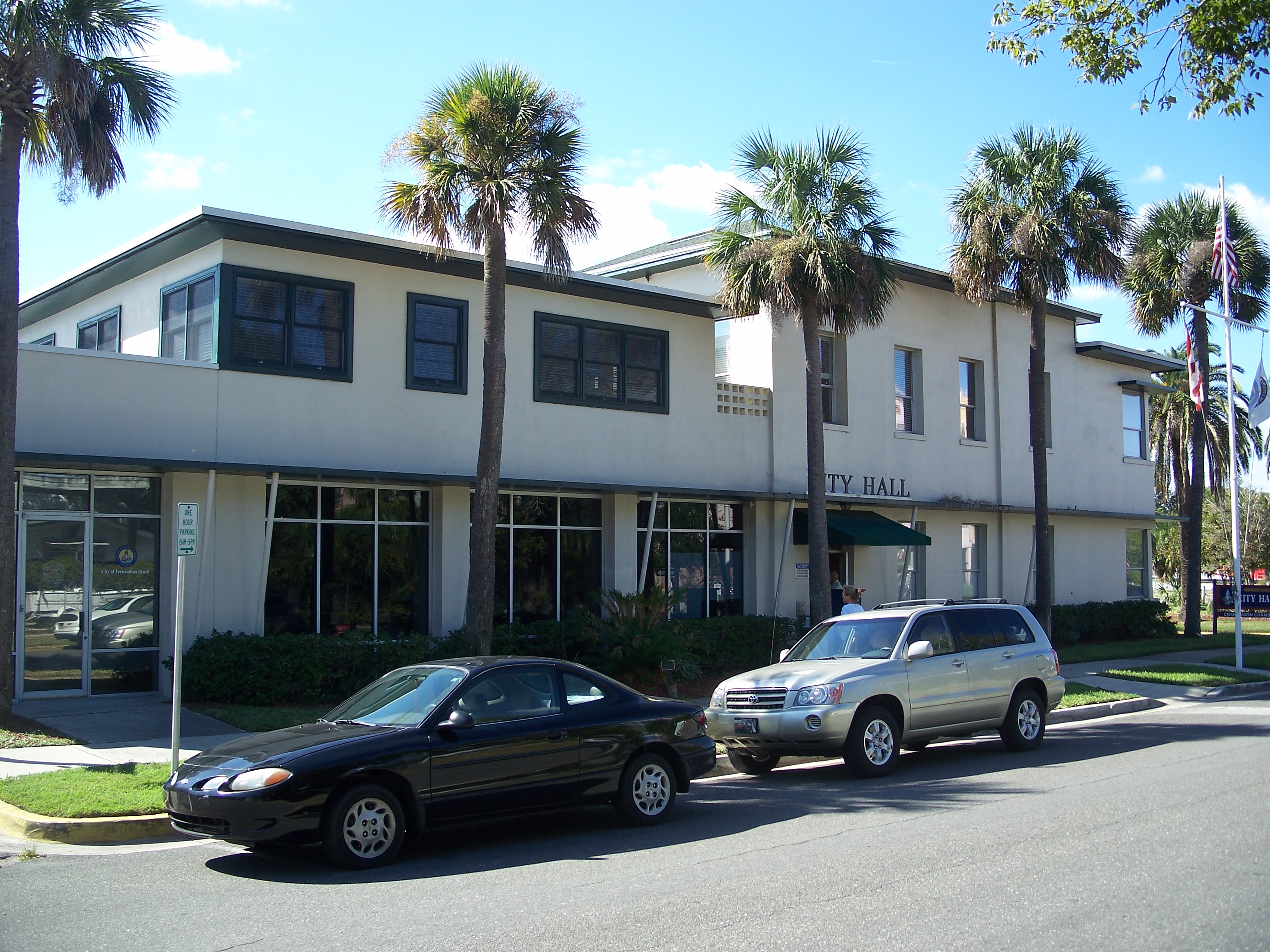 Front facade of a two-story beige city hall building with palm trees and flags.