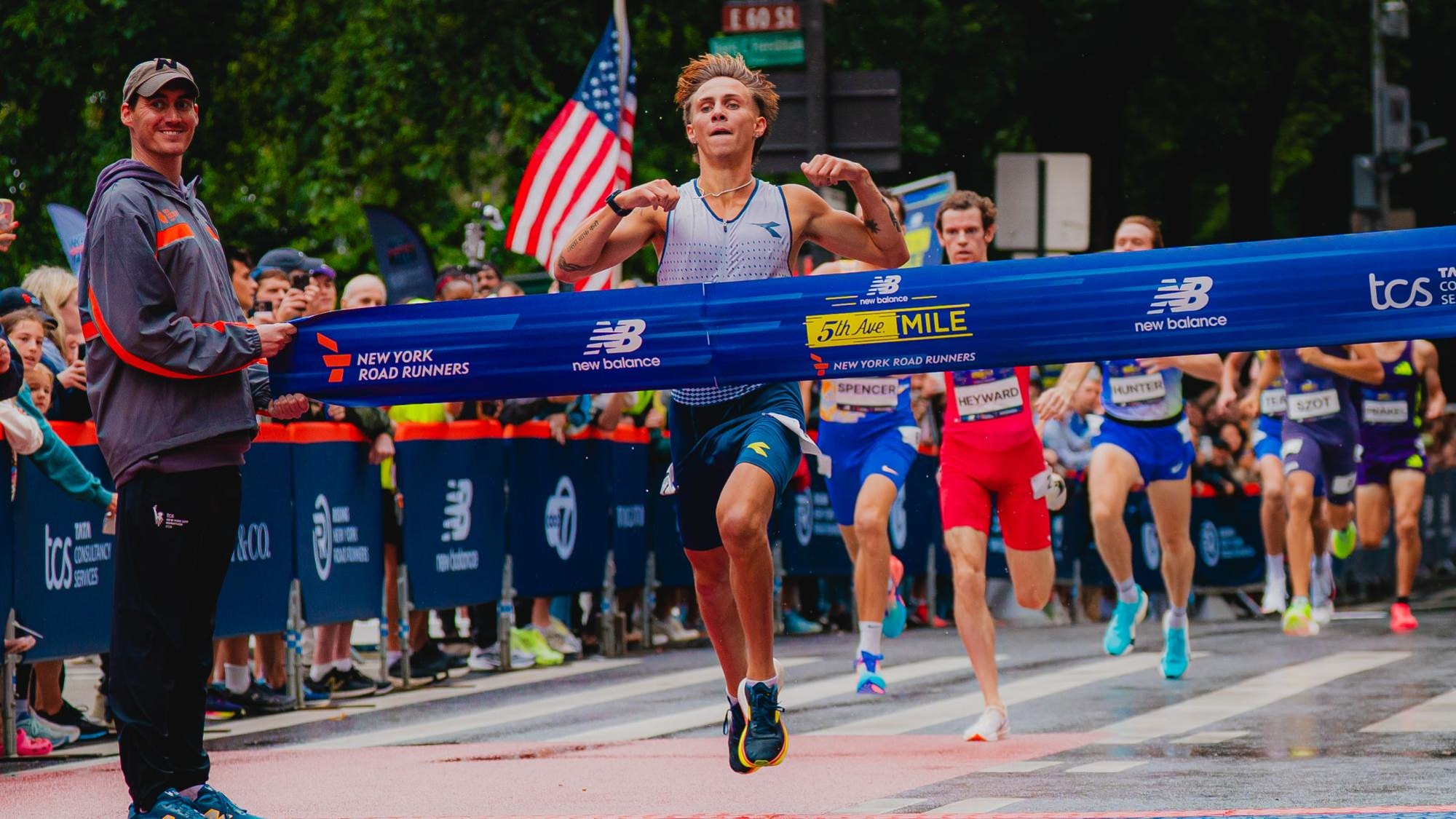 Male runner breaking the finish-line tape at a road mile race, with other runners close behind.
