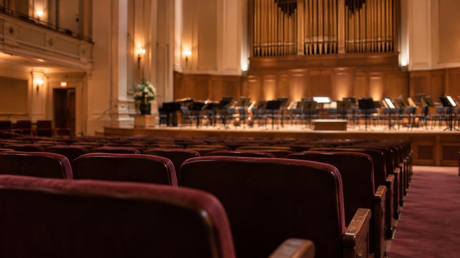 Empty concert hall auditorium with upholstered seats facing a warmly lit stage and pipe organ.