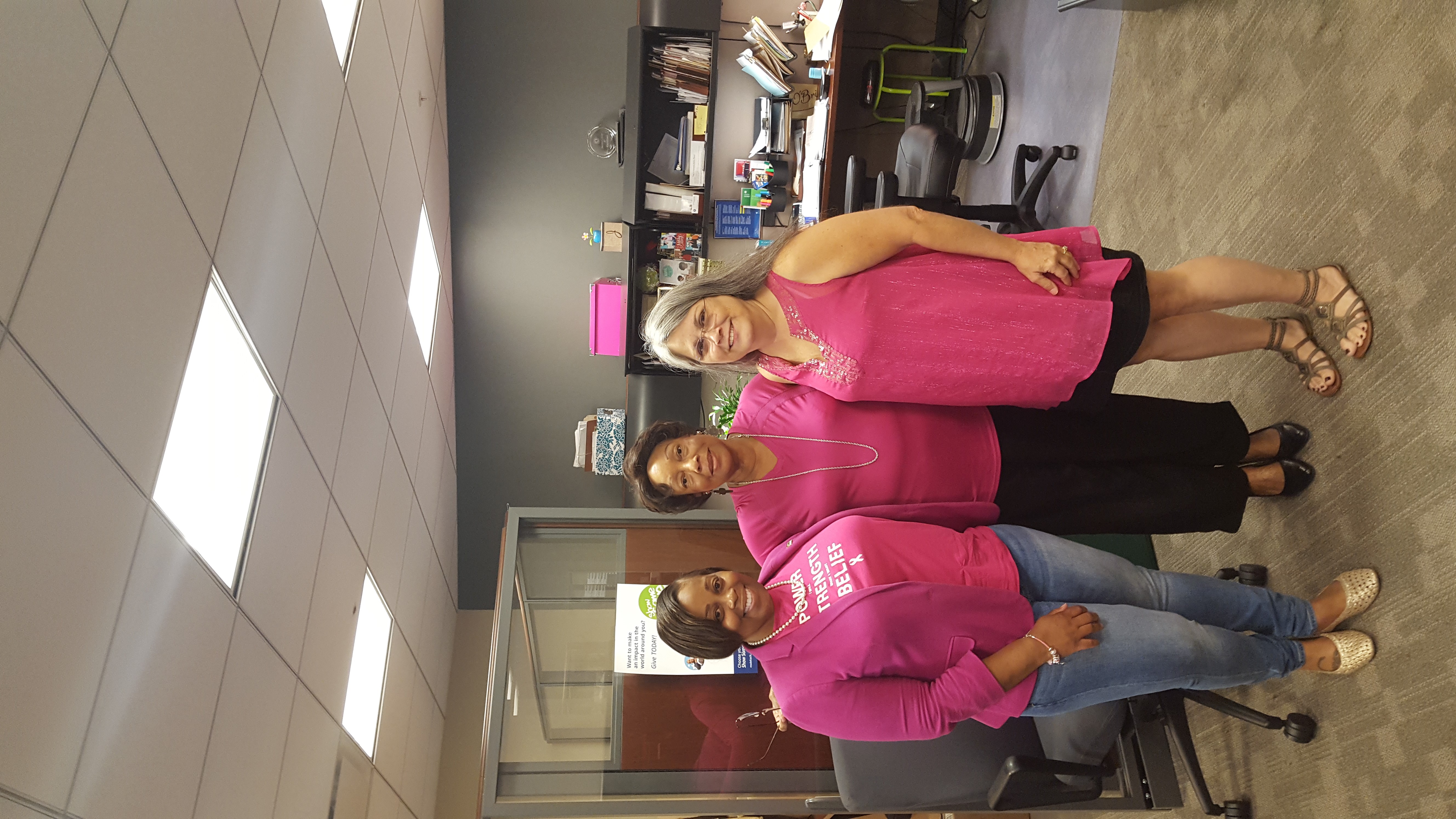 Three women wearing pink clothing stand together in an office setting.