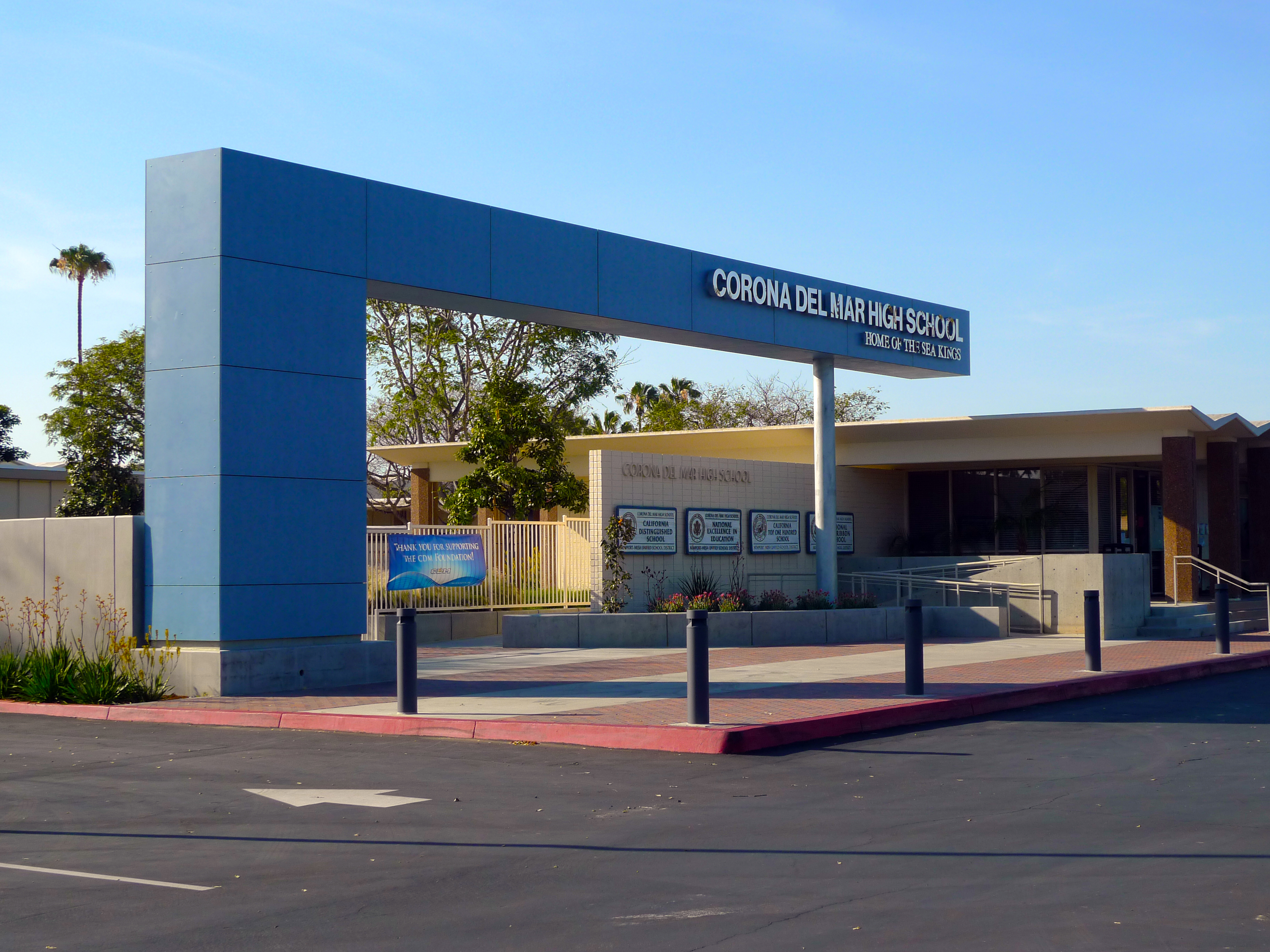 Blue entrance arch reading 'Corona del Mar High School' with palm trees and bollards.