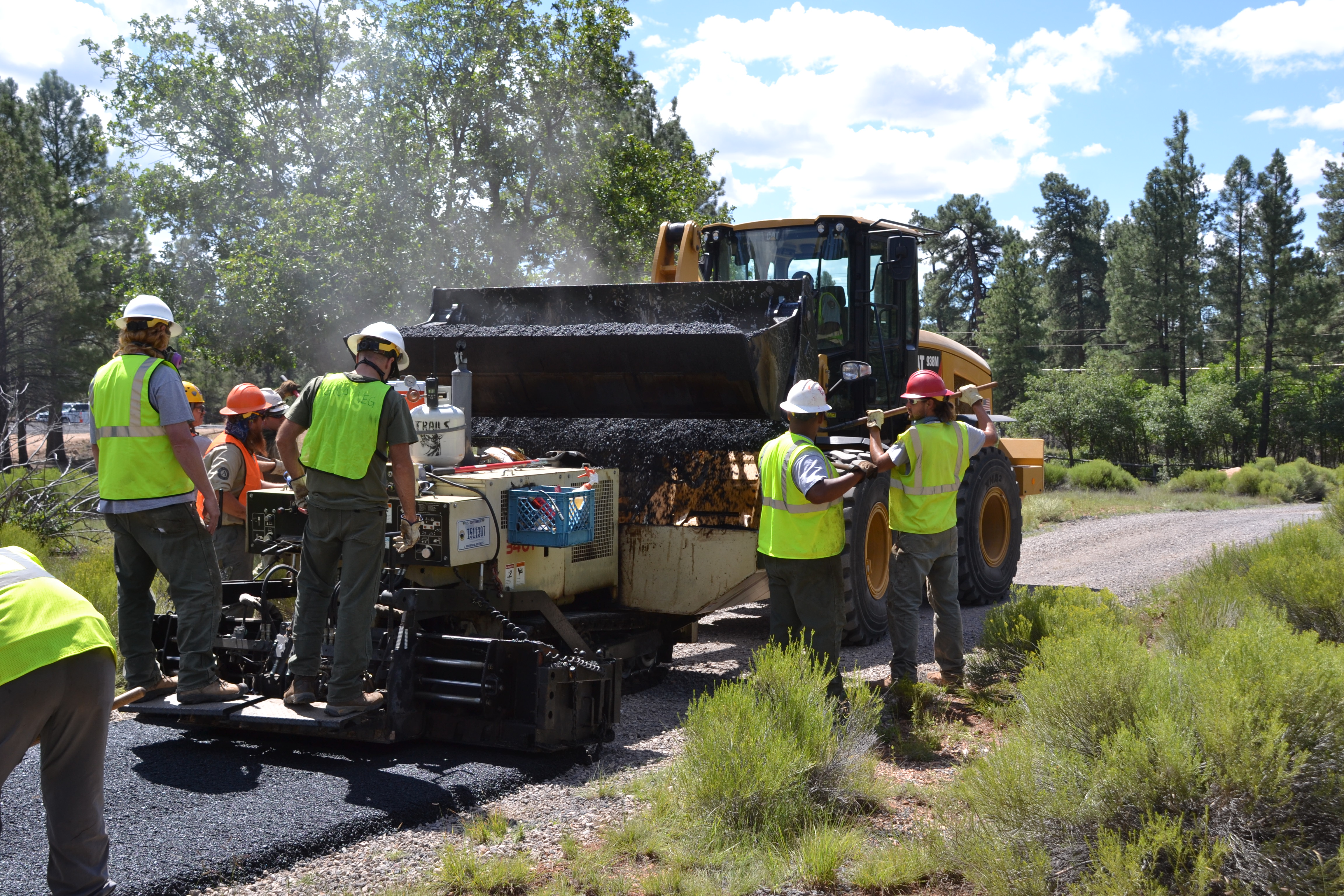 Workers in safety vests operate a paving machine laying asphalt on a road.