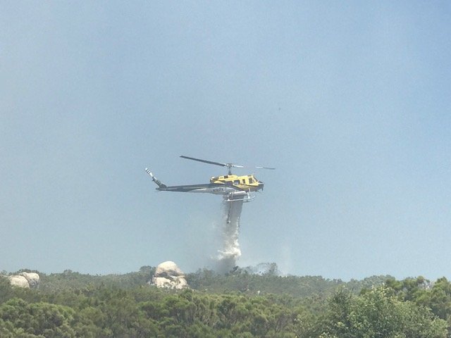 Yellow and white helicopter releasing water over a smoke-hazed shrubby hillside.