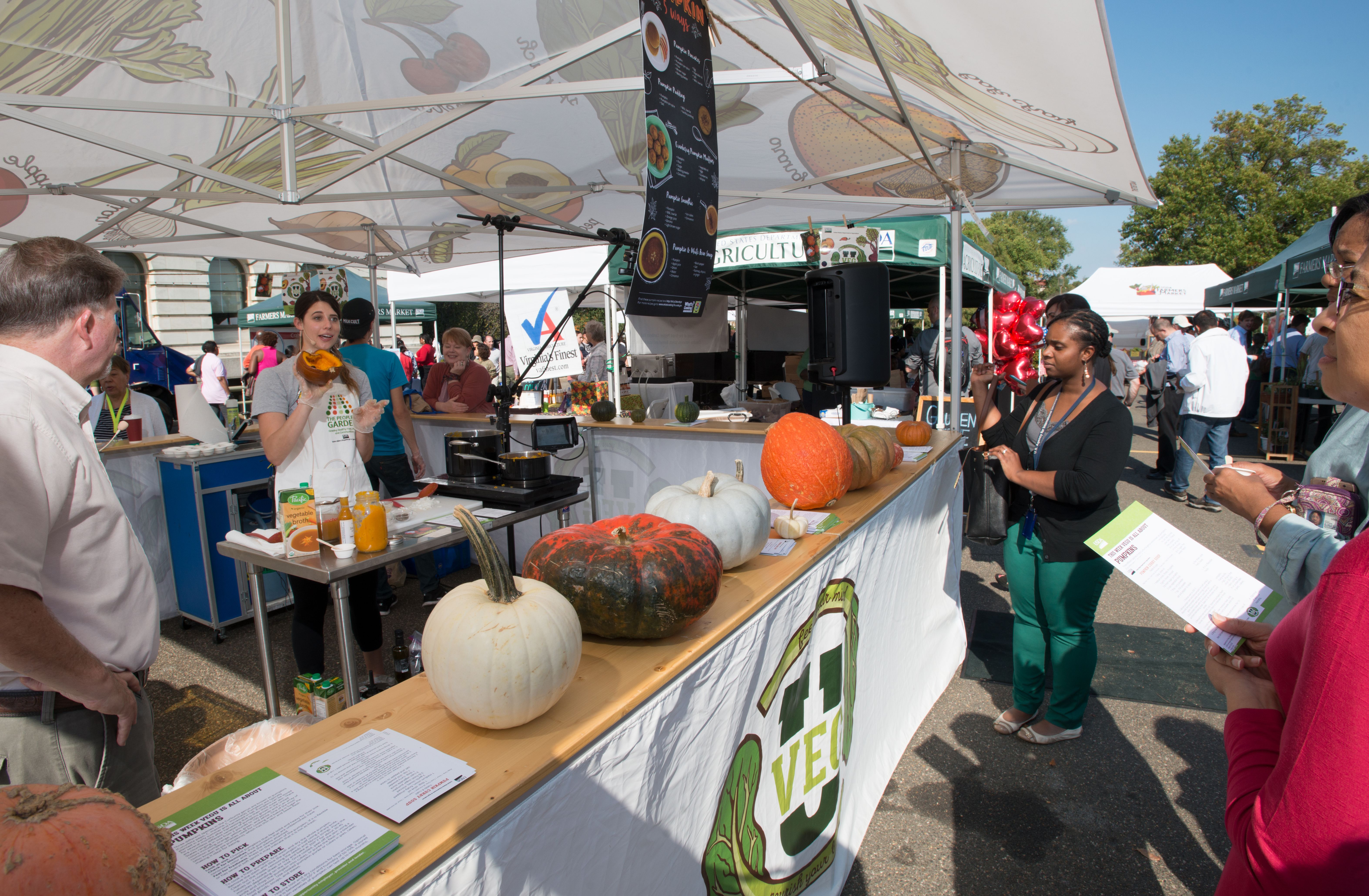 Outdoor farmers market booth with pumpkins and gourds under a white canopy tent.