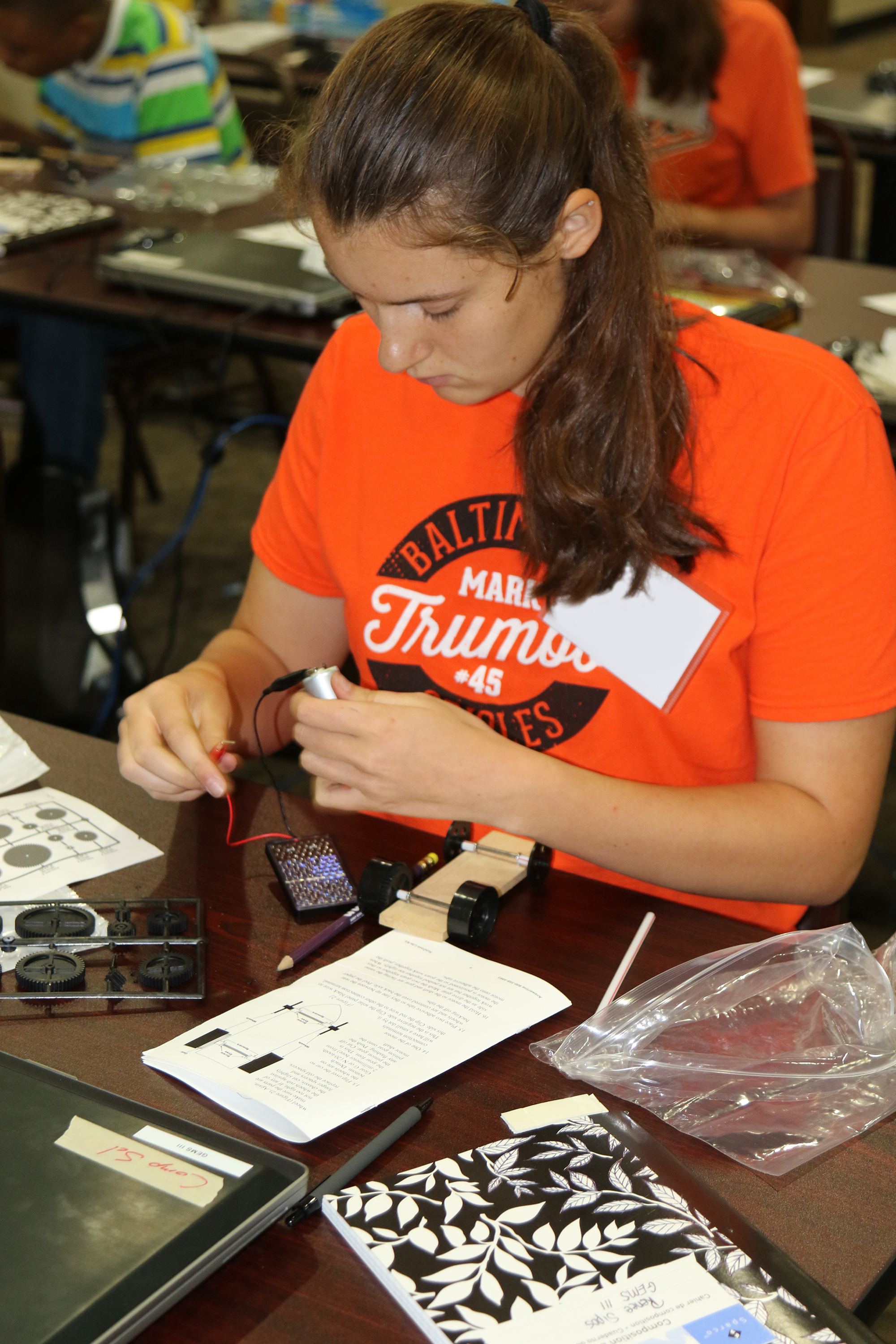 A student in an orange shirt works with electronic components at a classroom desk.
