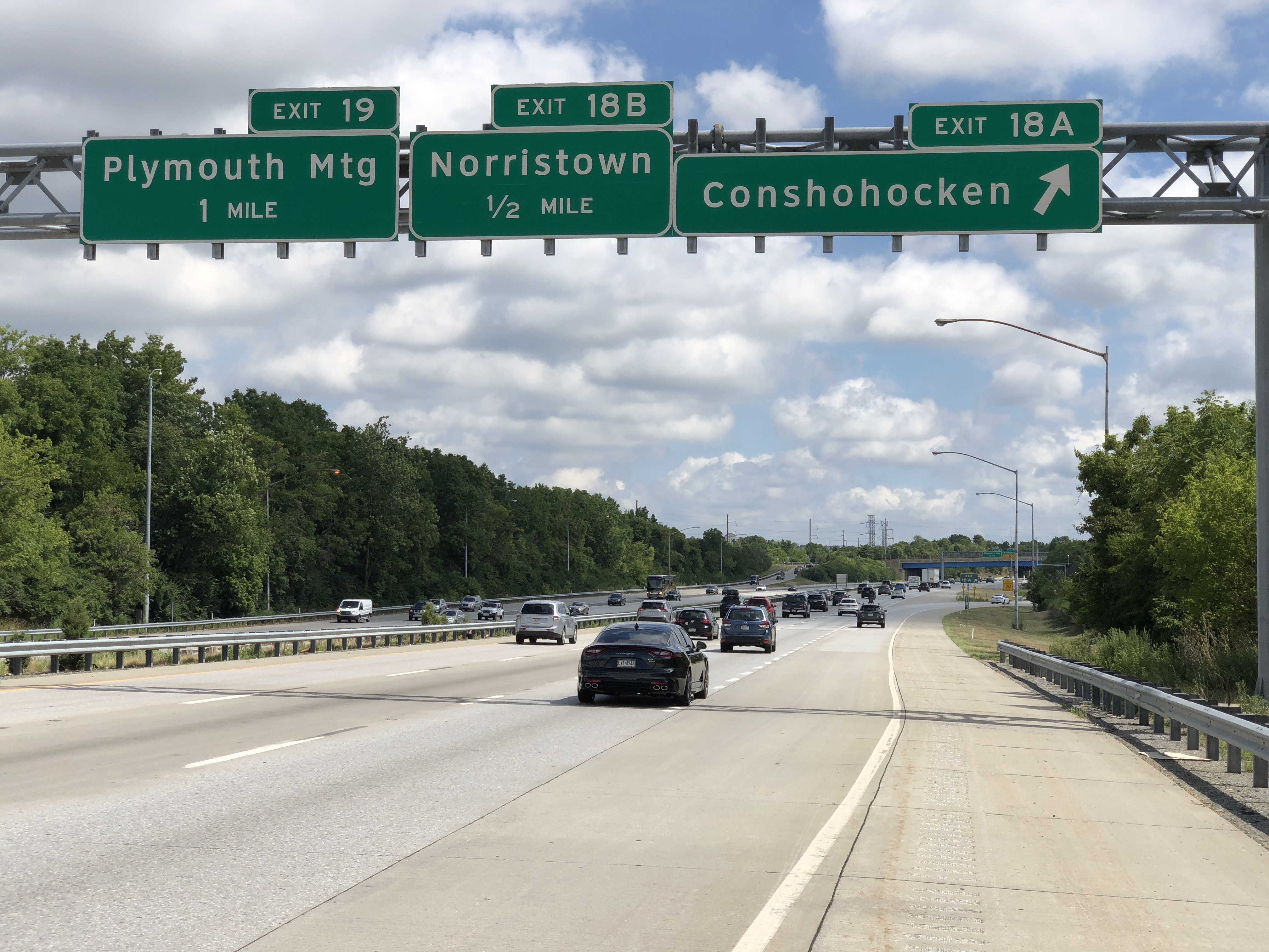 Northbound highway lanes with overhead green signs for Conshohocken and Plymouth Meeting exits.