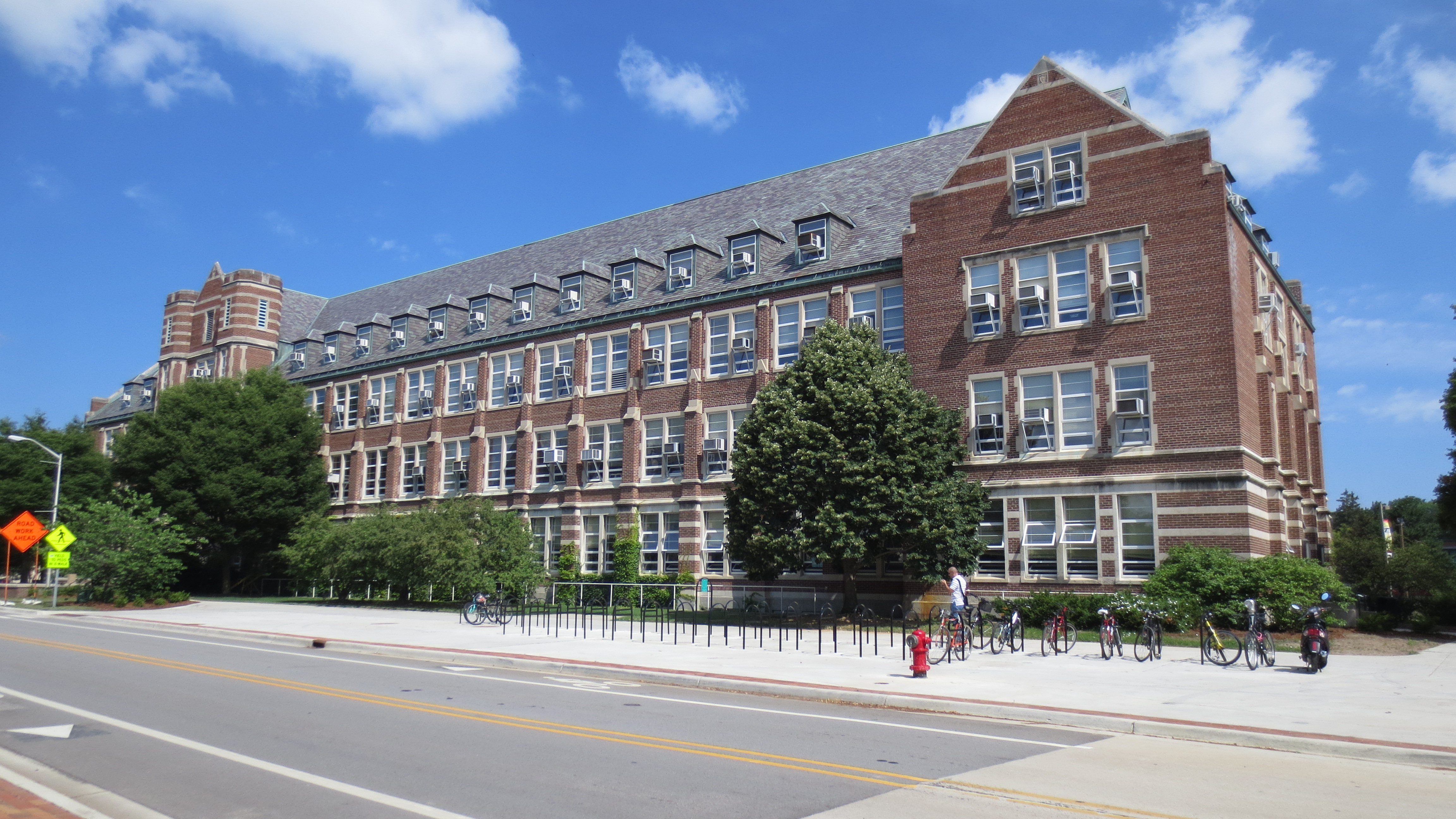 Exterior of a large red brick university building with dormer windows and parked bicycles.