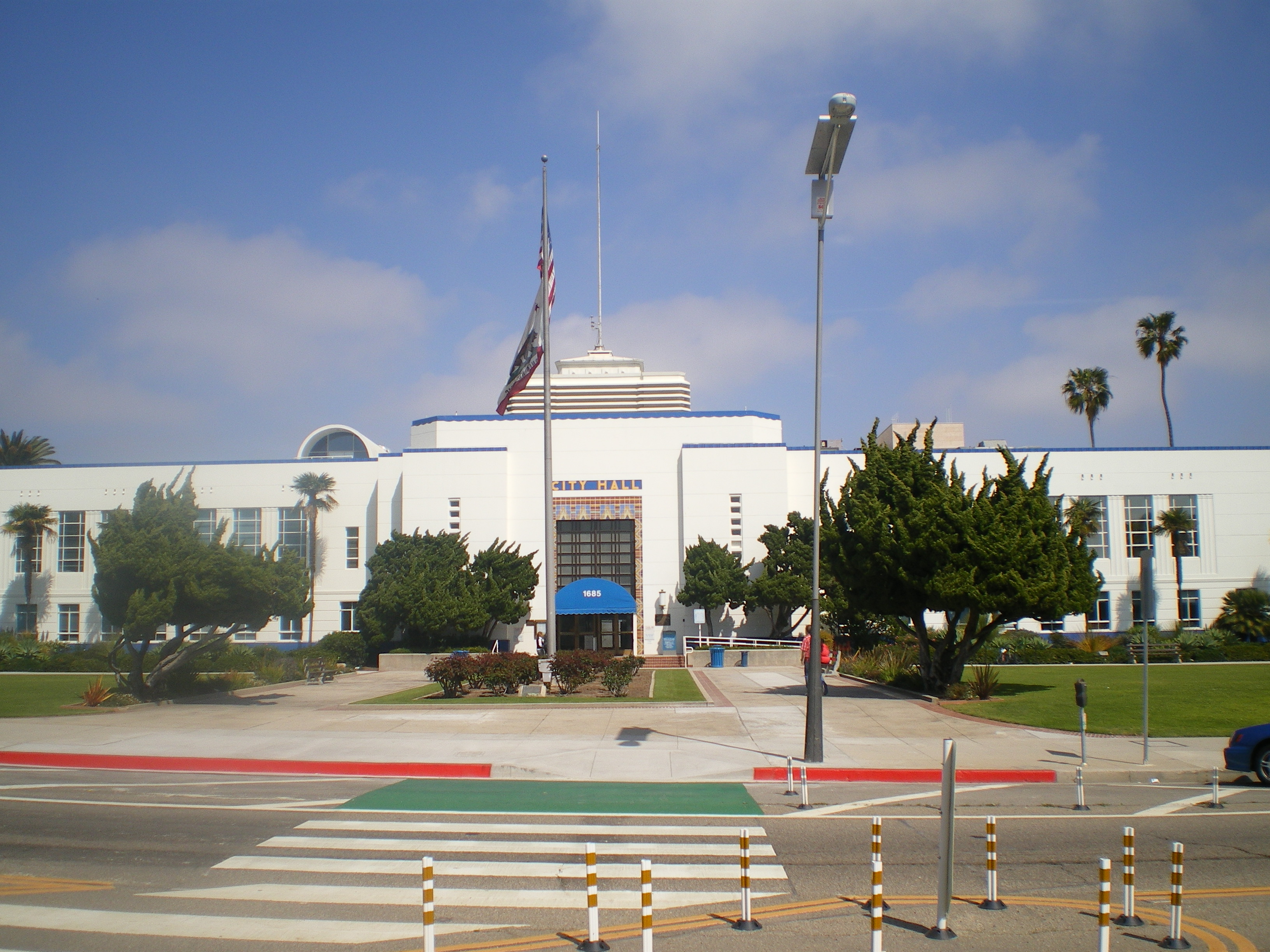 White Art Deco city hall building with an American flag and palm trees, viewed from across the street.