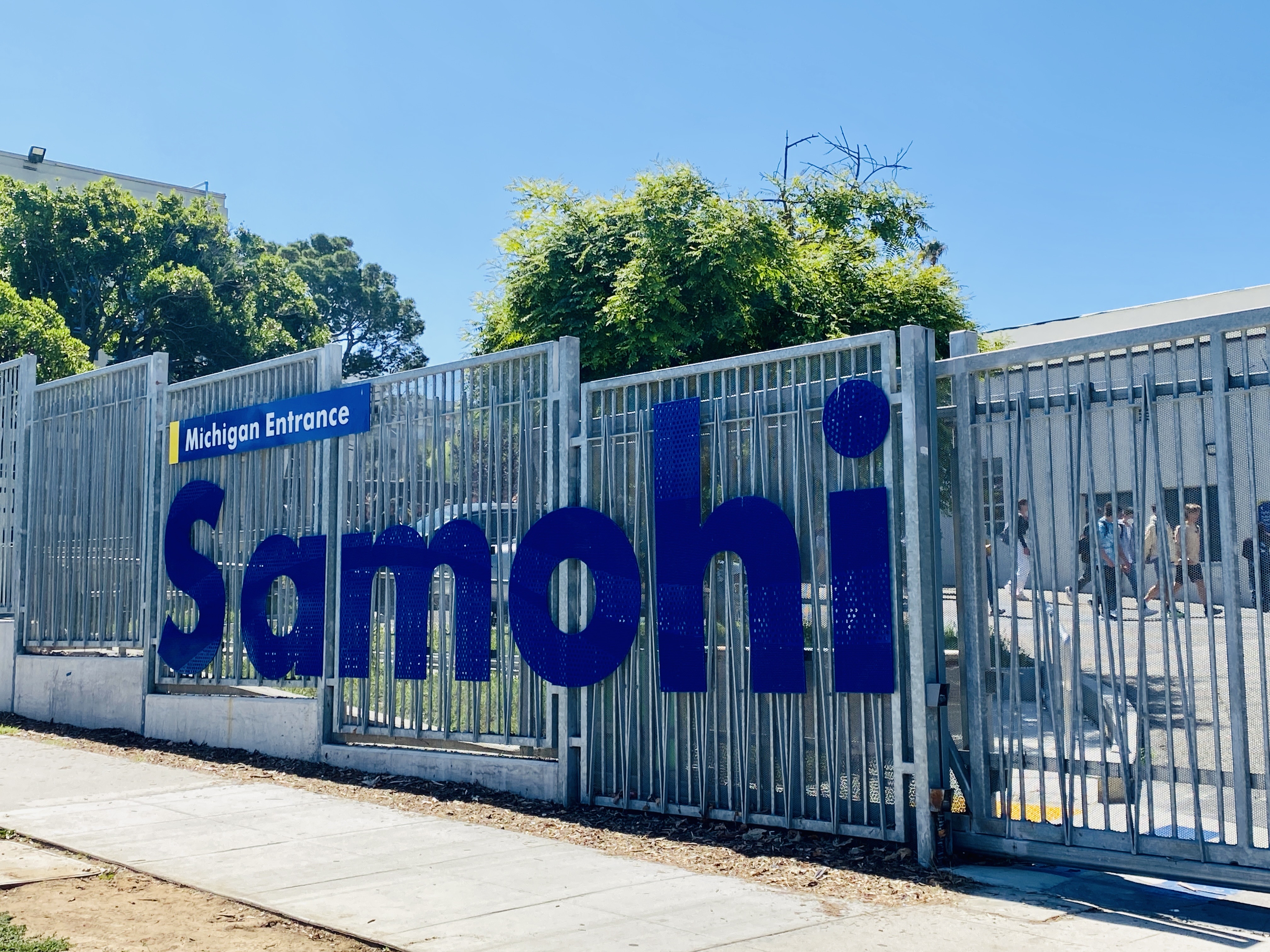 White metal school entrance gate with blue 'Samohi' lettering and trees behind it.