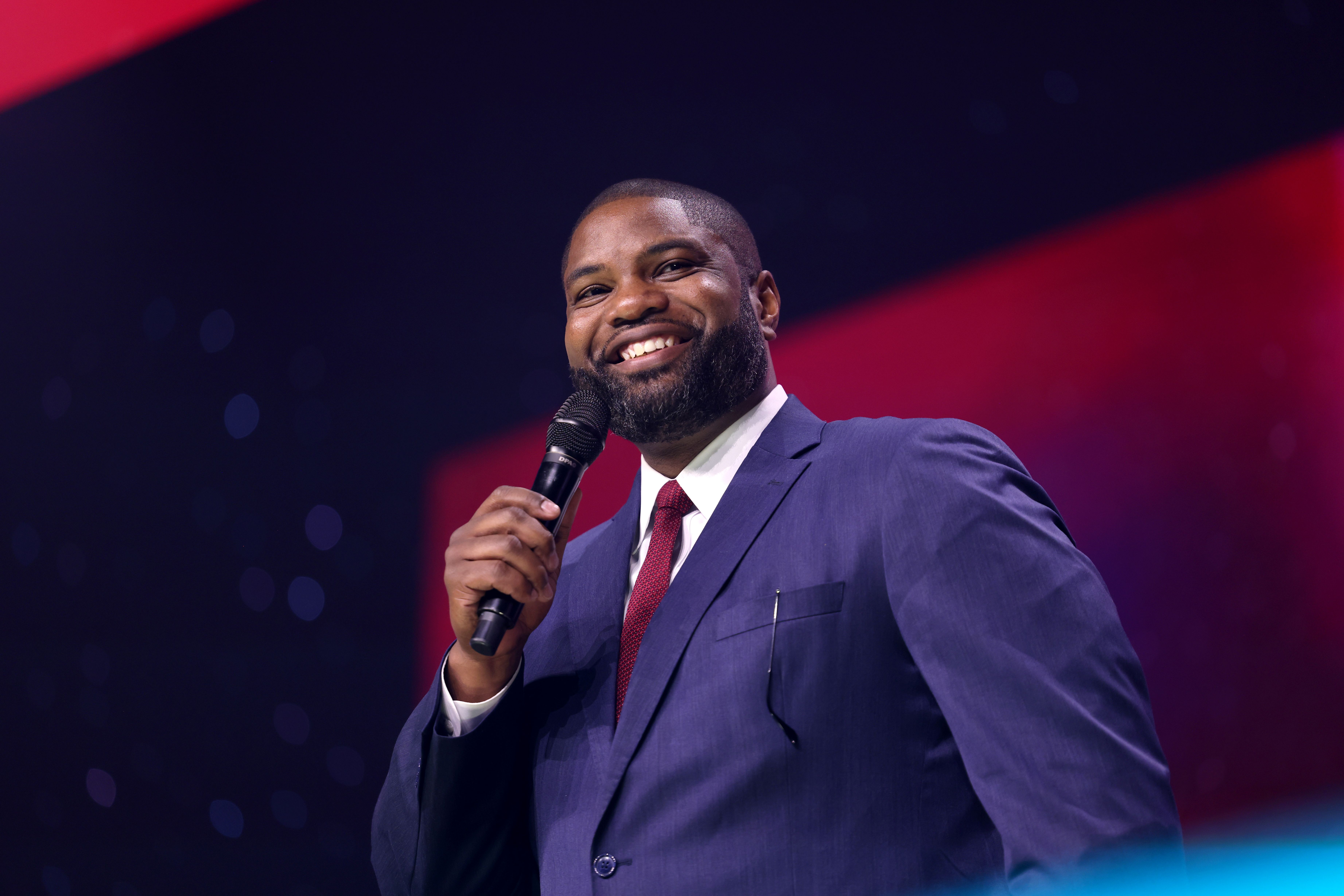 A man in a blue suit smiles and holds a microphone on a stage.