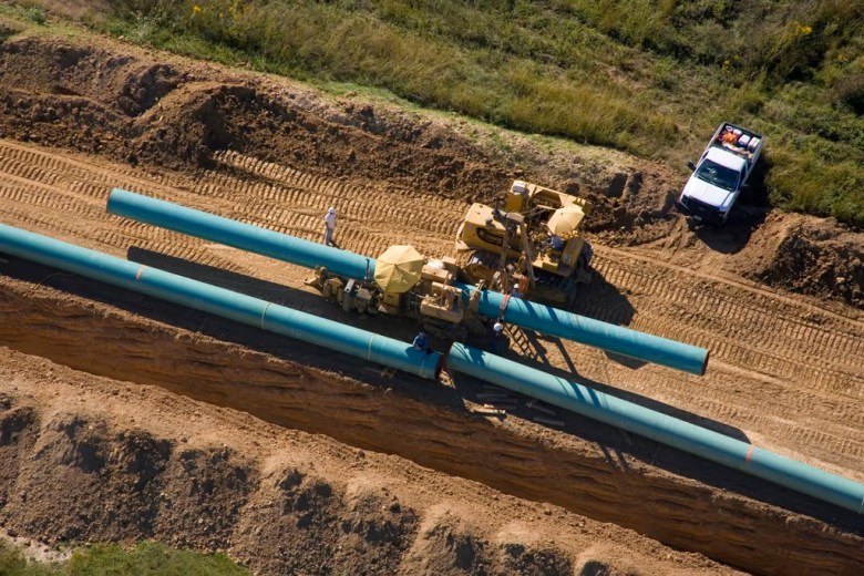 Aerial view of large blue pipes laid in a trench with yellow construction equipment nearby.
