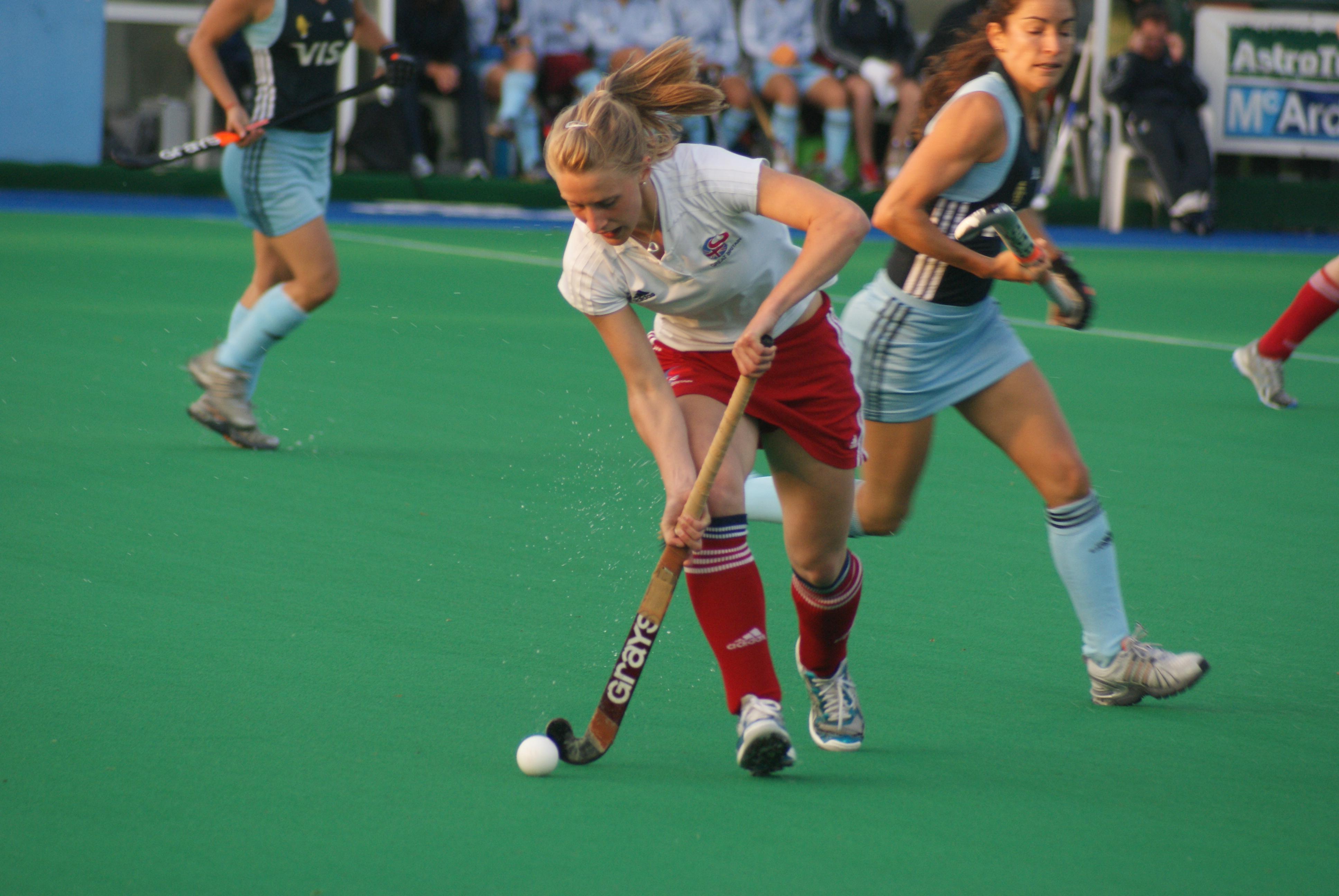 Female field hockey players competing for the ball on a green turf field.