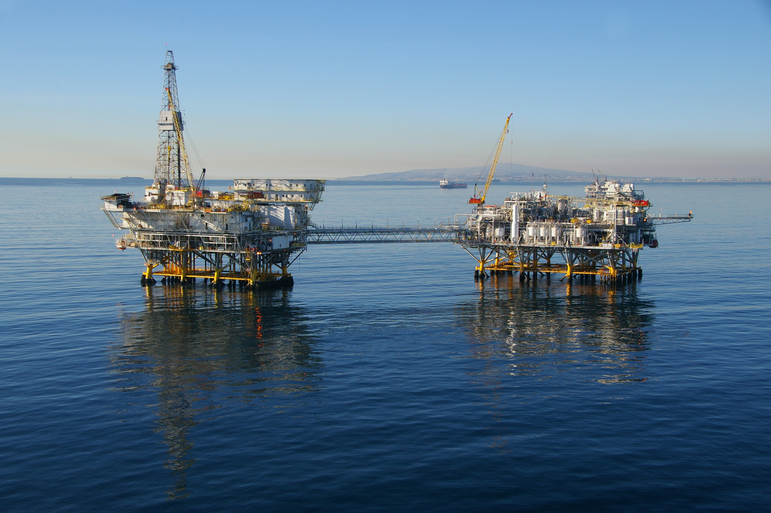 Two offshore oil drilling platforms on calm blue water with mountains in the background.