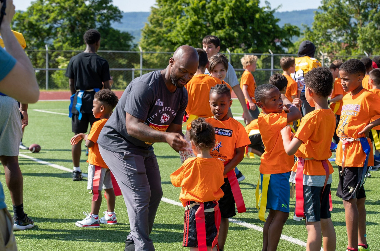 An adult coach bends down to talk with young children in orange shirts on a football field.