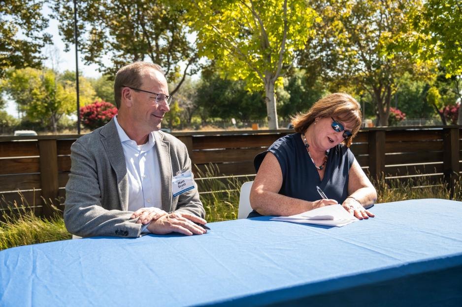 Two people seated at an outdoor table, one signing a document while the other watches.