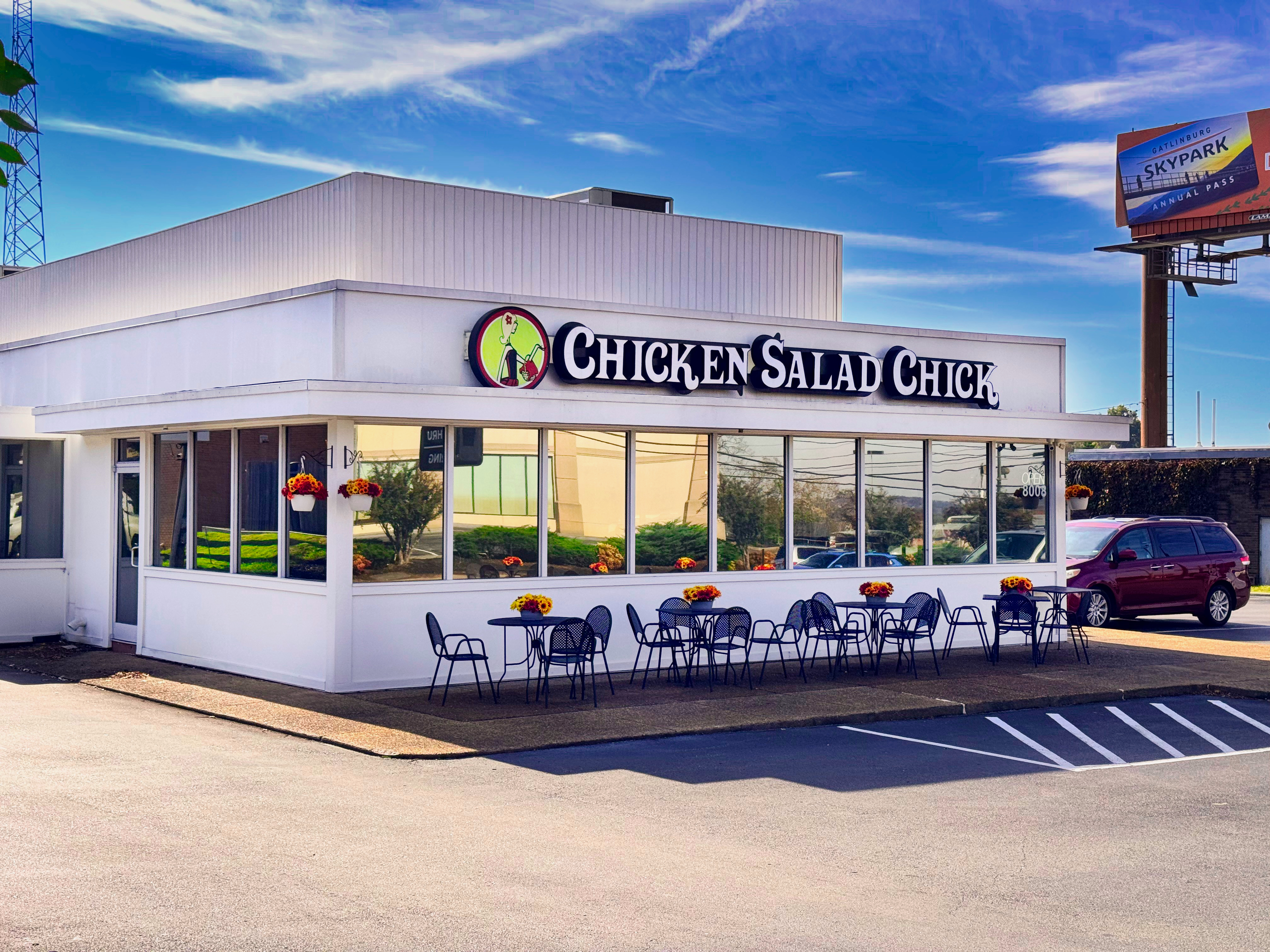 Exterior of a white Chicken Salad Chick restaurant with outdoor seating and flower baskets.