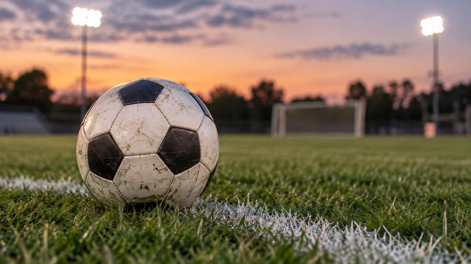 A soccer ball on a grass field with stadium floodlights glowing in the background at dusk.