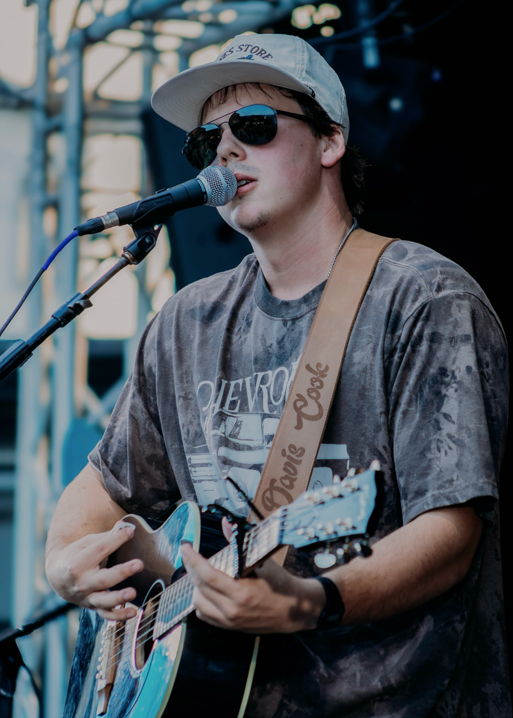 Young man in a backward cap plays acoustic guitar and sings into a microphone on an outdoor stage.