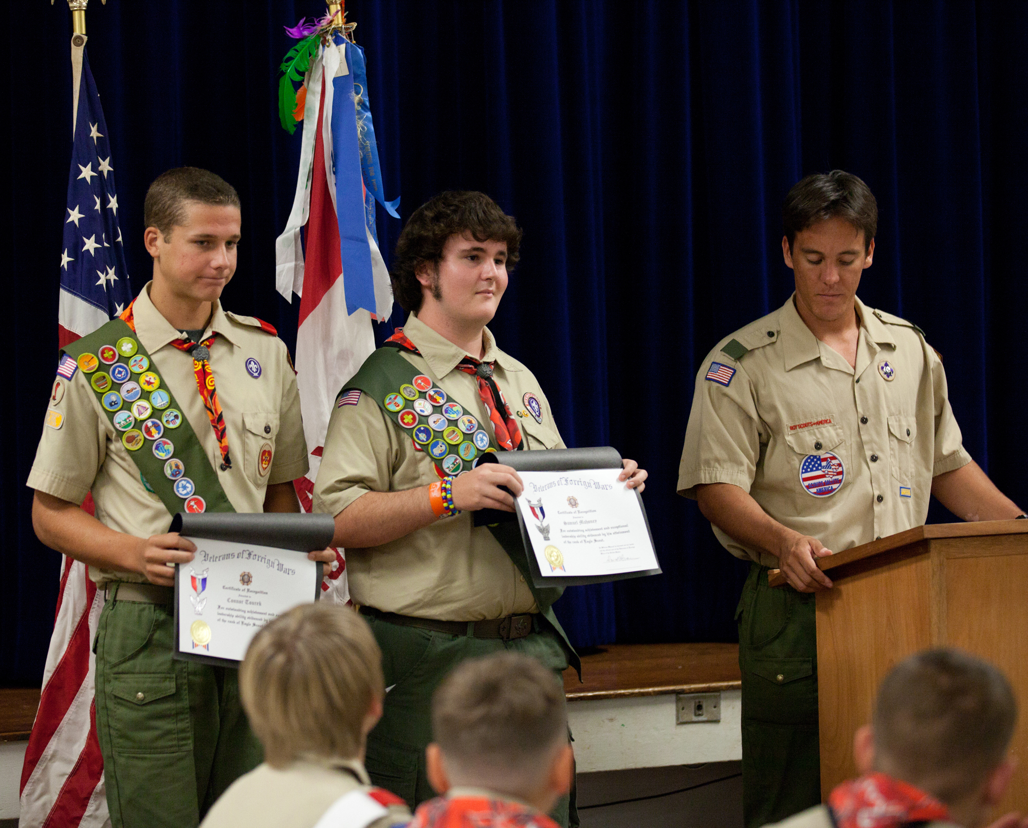 Two scouts in tan uniforms with merit badge sashes hold certificates at a ceremony.