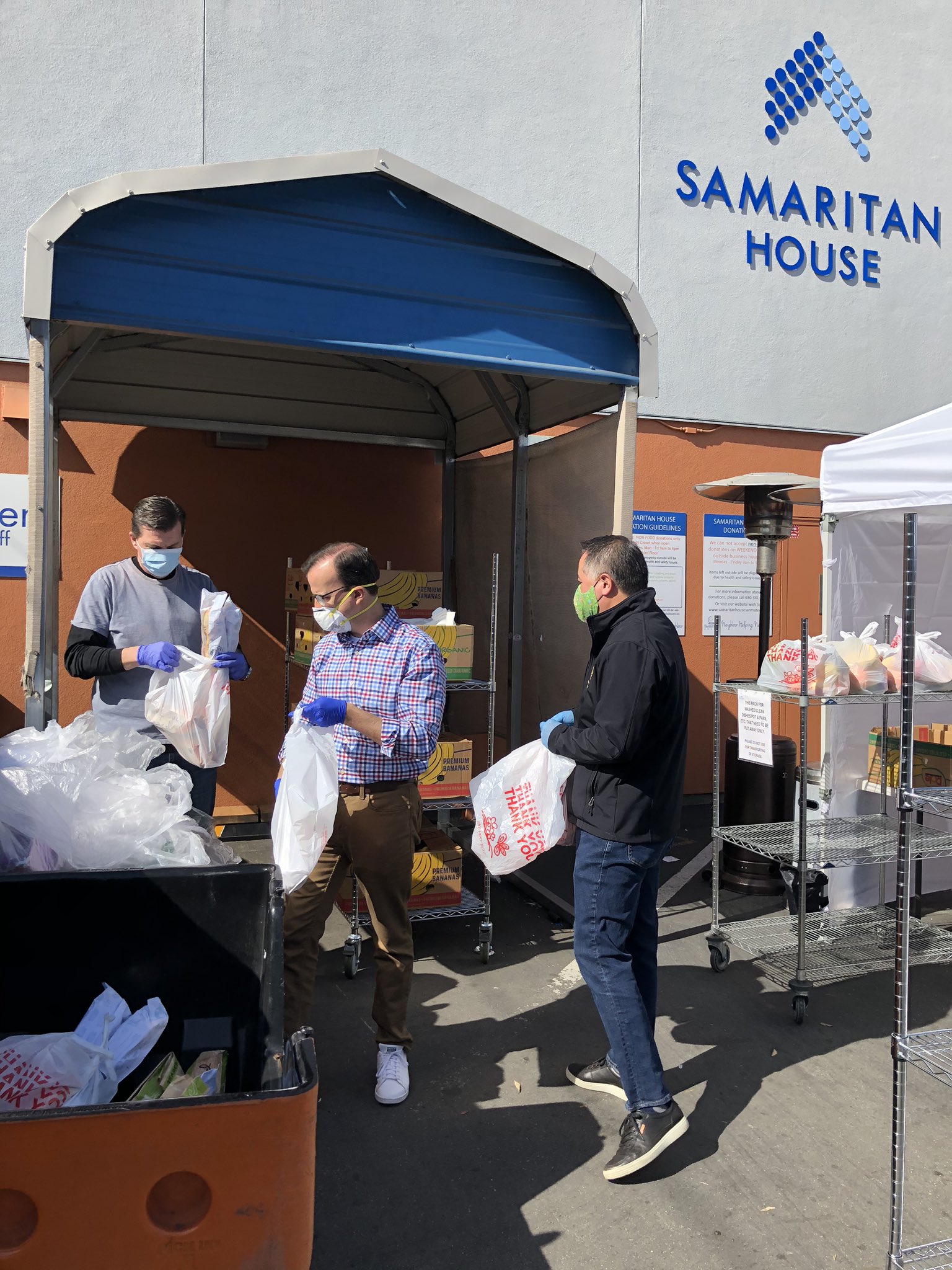 Three masked volunteers hand out bags of supplies under an awning at Samaritan House.