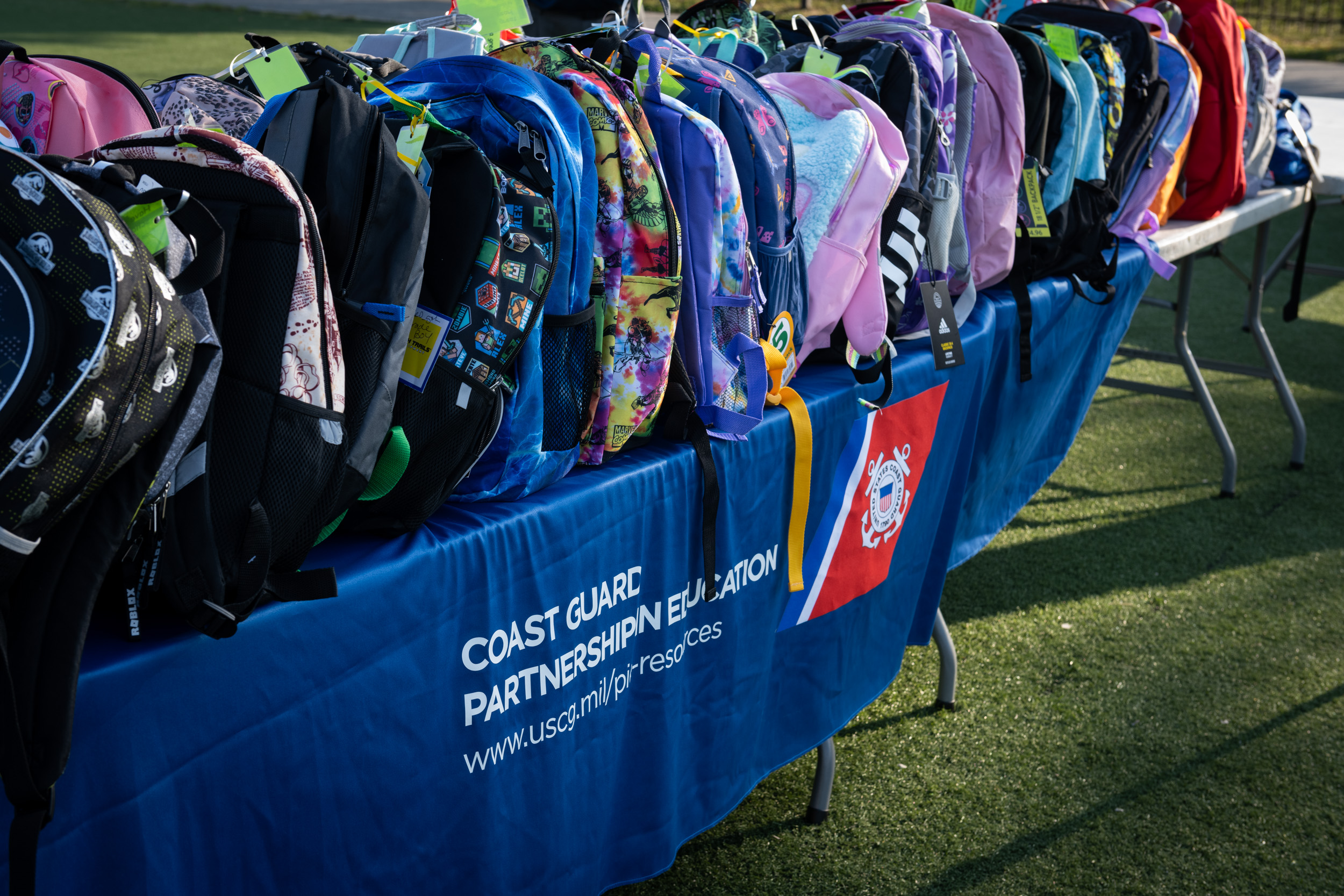 A long table covered with a blue banner holds dozens of colorful backpacks of various sizes outdoors.