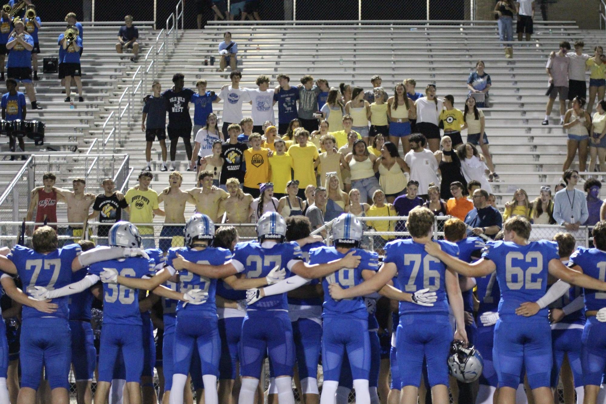 Football players in blue uniforms stand arm-in-arm facing a crowded student section at night.