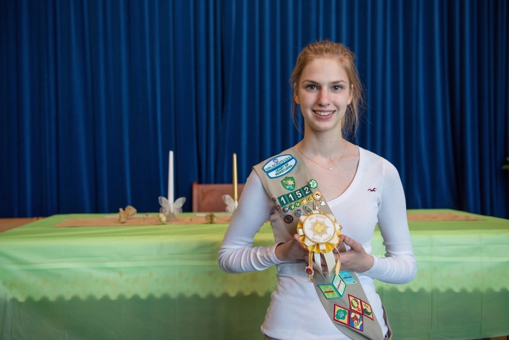Smiling young woman in a Girl Scout sash holding a Gold Award rosette at a ceremony.