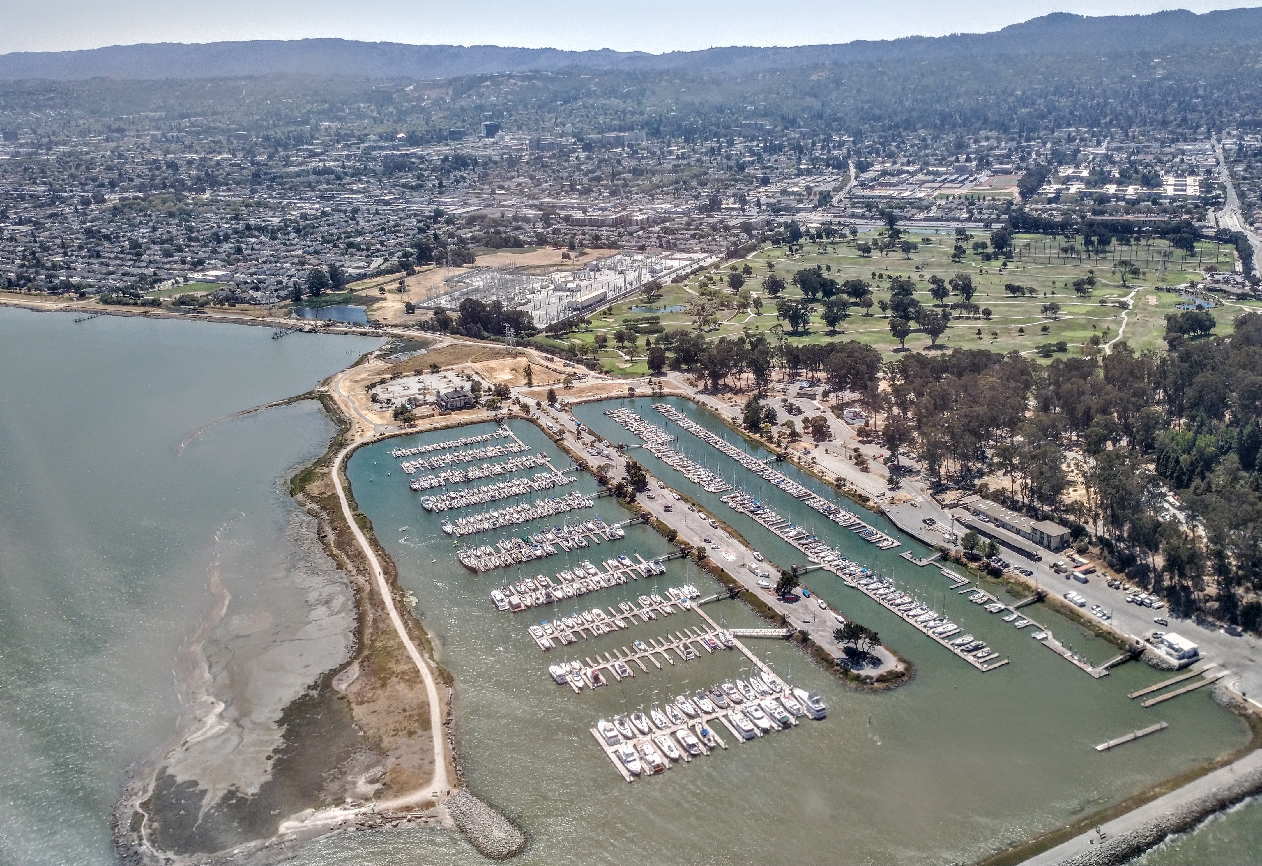 Aerial view of a waterfront park with a marina, green lawns, and suburban city beyond.