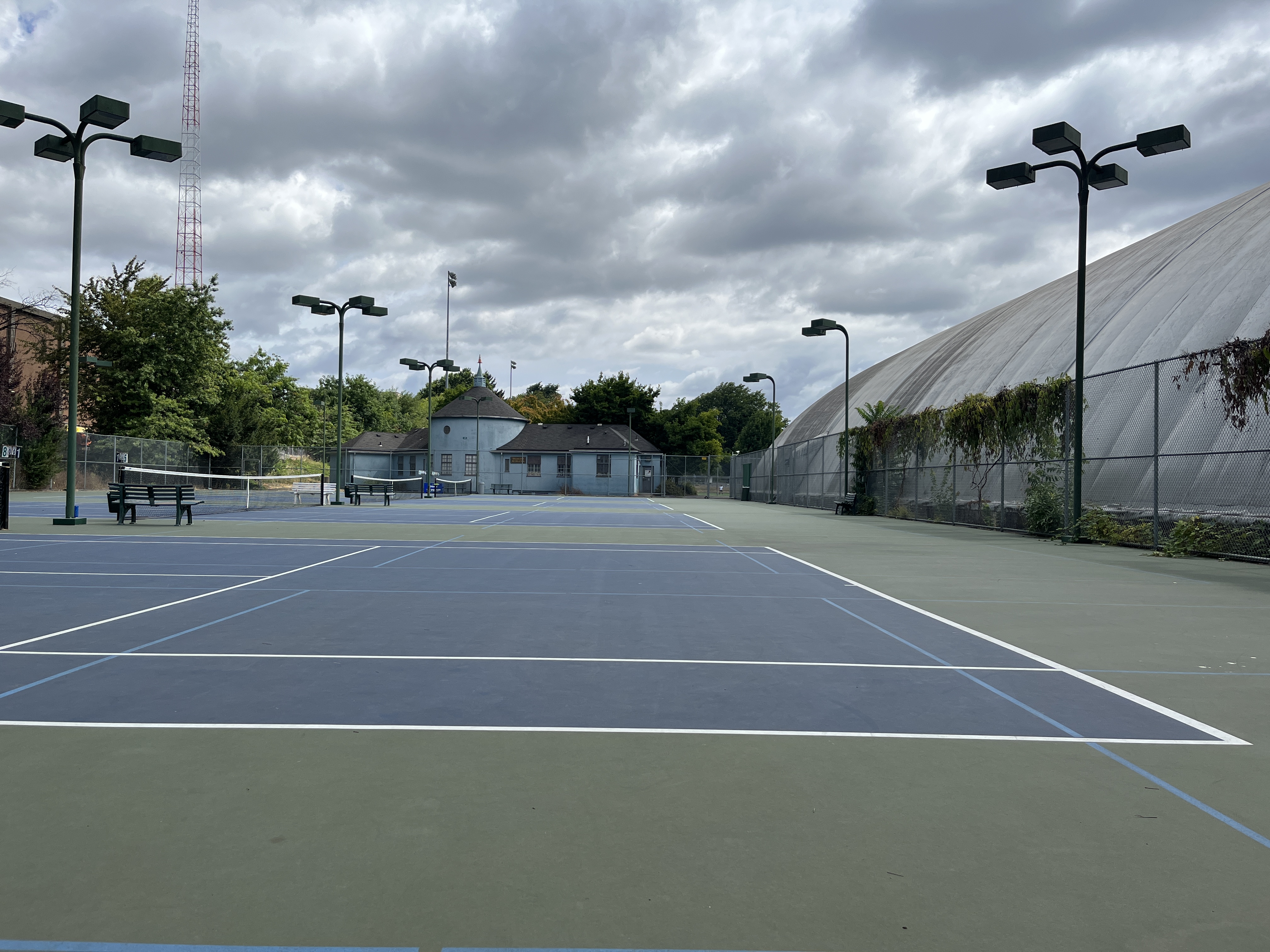 Outdoor blue tennis courts with chain-link fencing and tall floodlight poles.