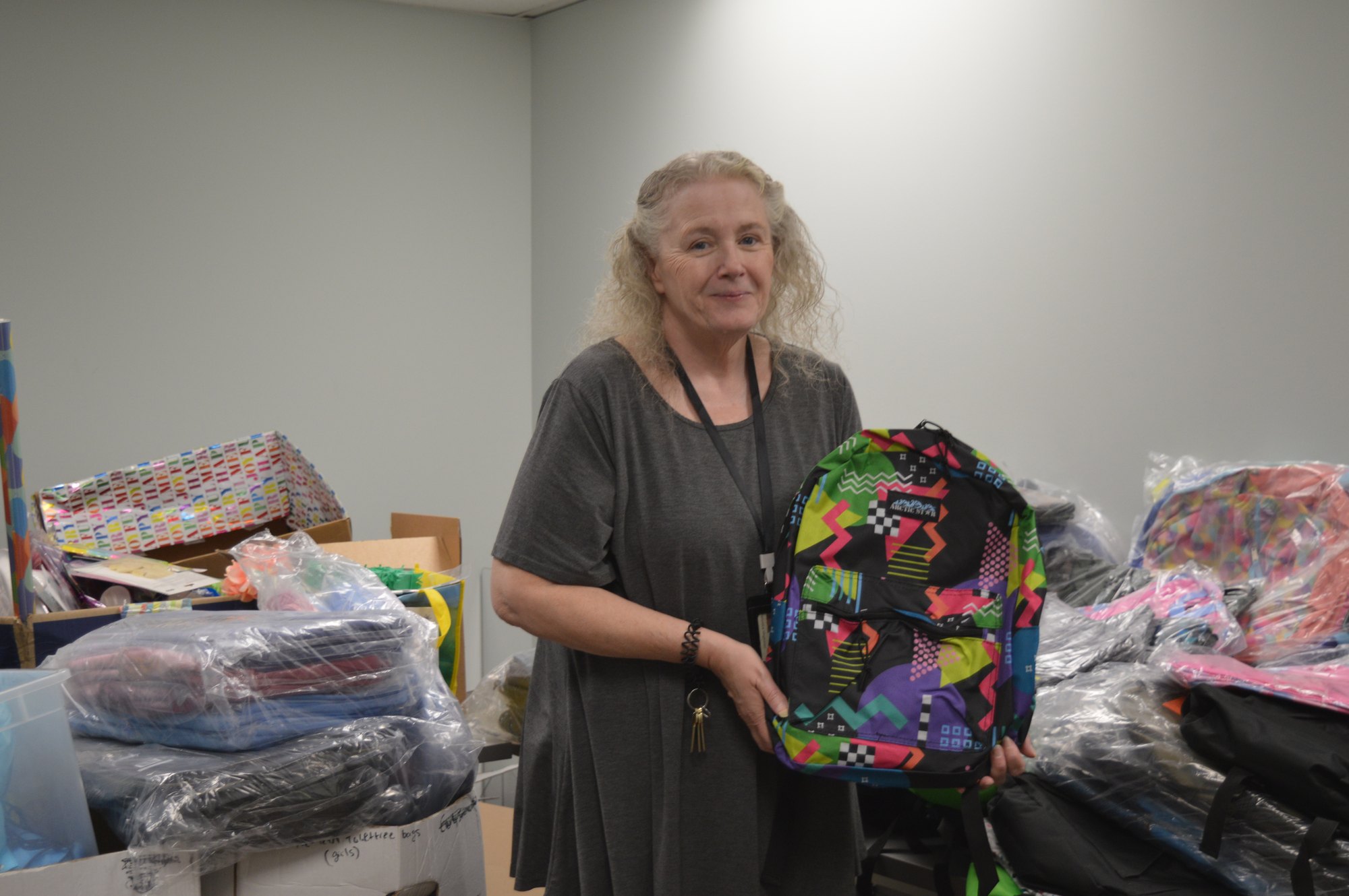 A woman in a gray dress holds a colorful patterned backpack amid bags of donated supplies.