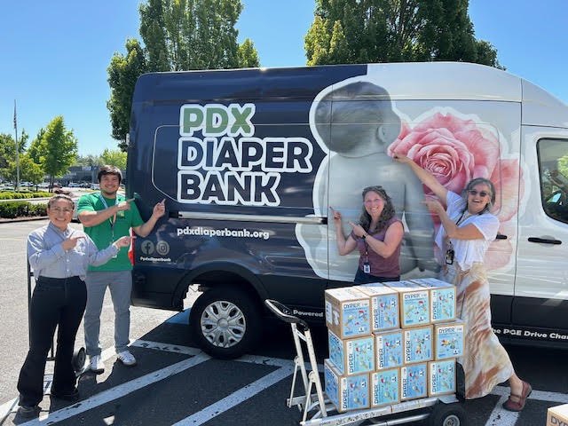 Four people pose beside a branded white van next to a pallet of boxed diapers.