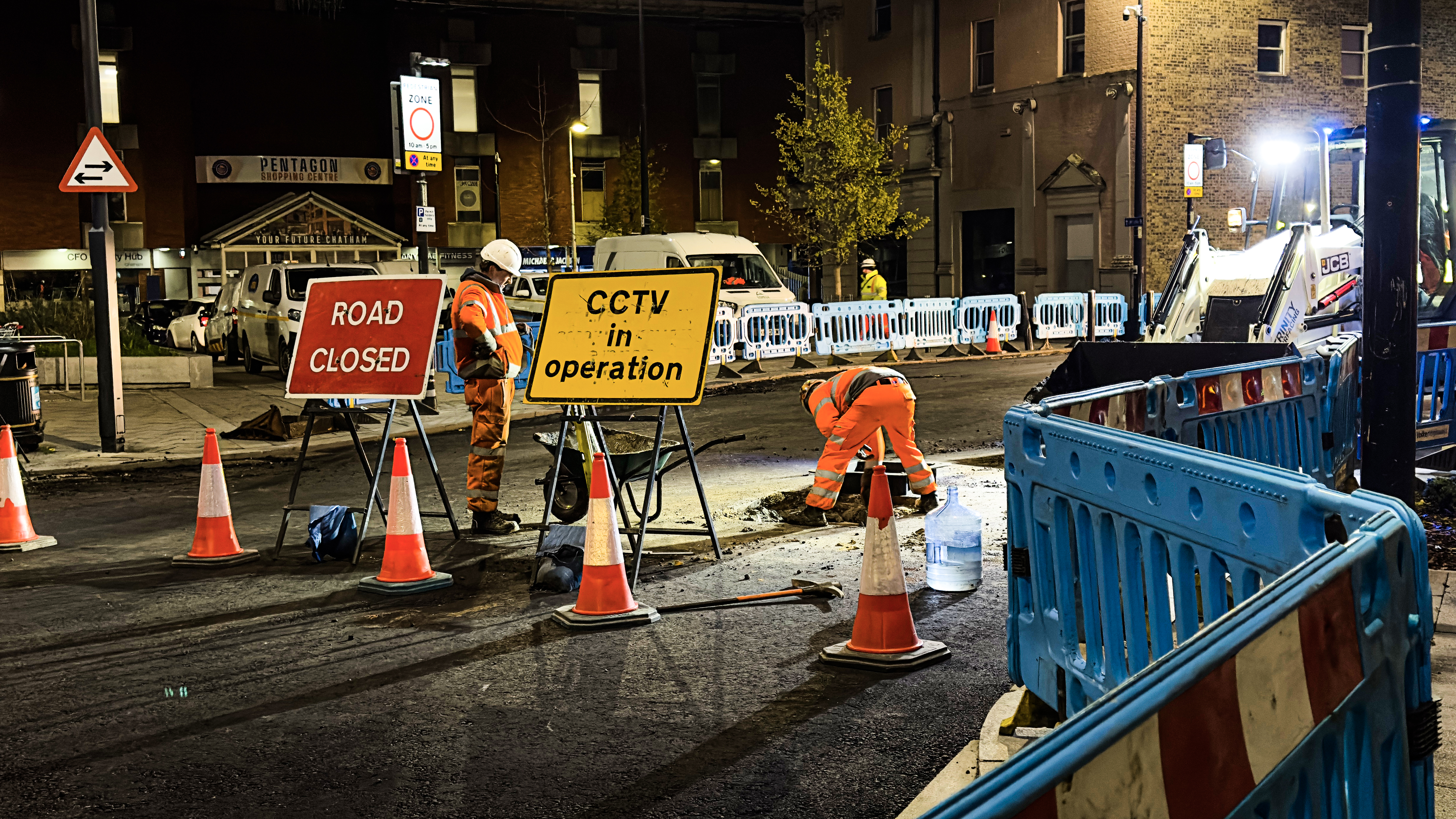 Two workers in orange vests at a nighttime road closure with cones and barriers.