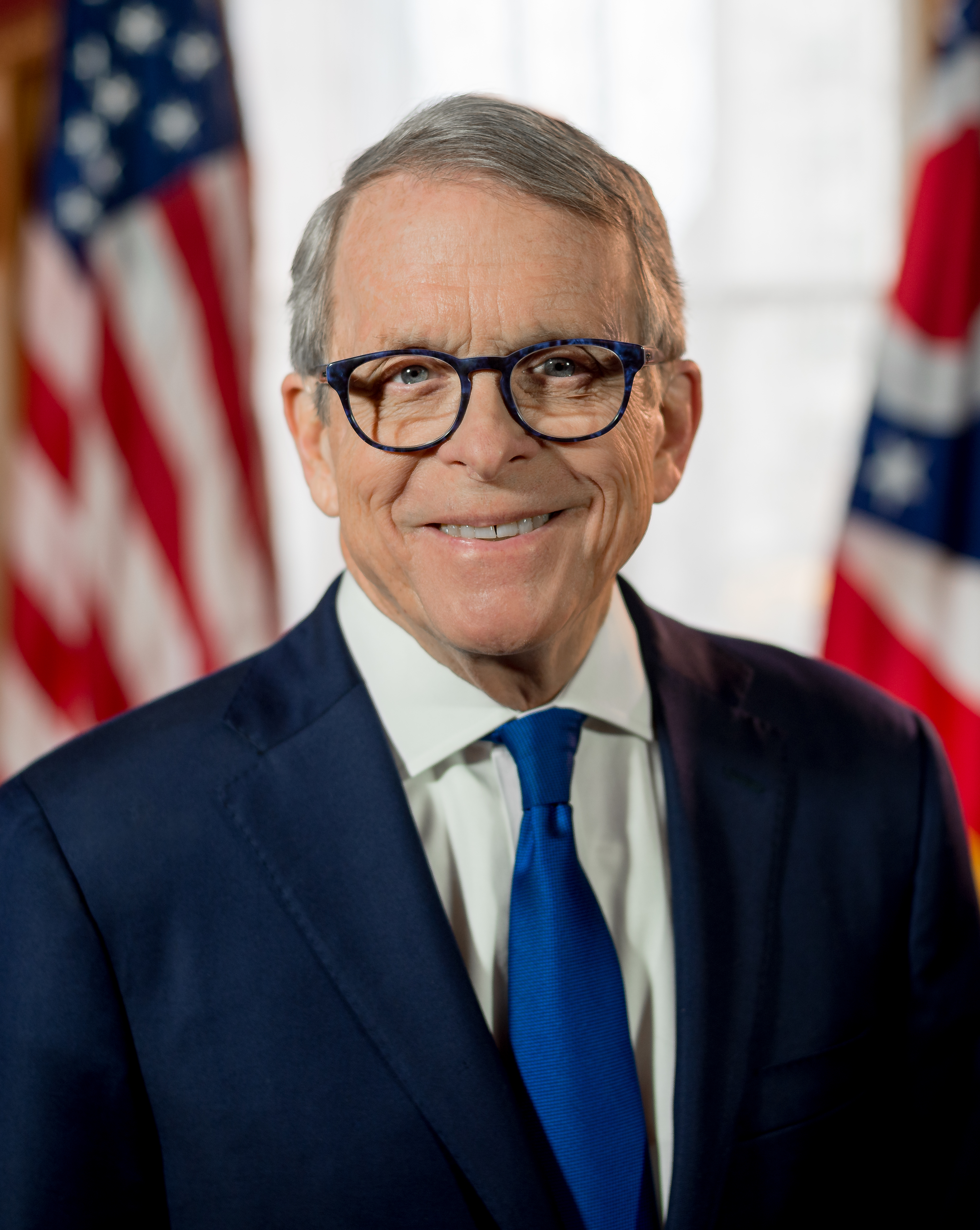 Man in dark suit and blue tie smiling before American and Ohio state flags.