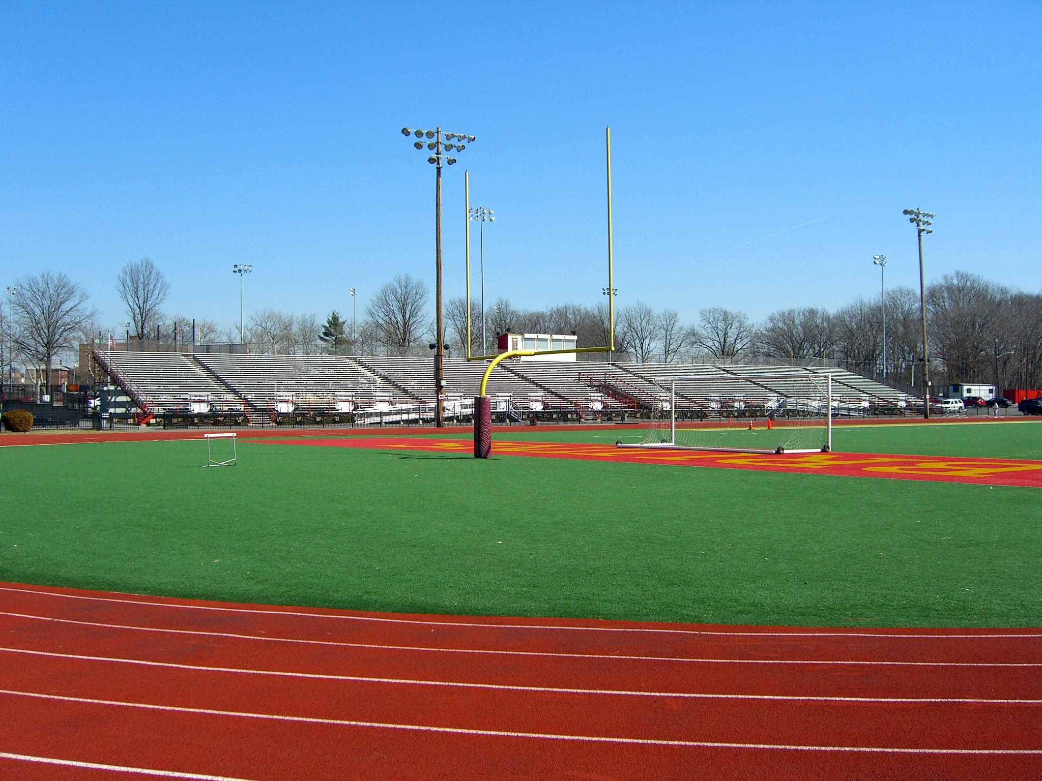 Empty high school football stadium with red track, green turf, yellow goalposts, and stadium lights.