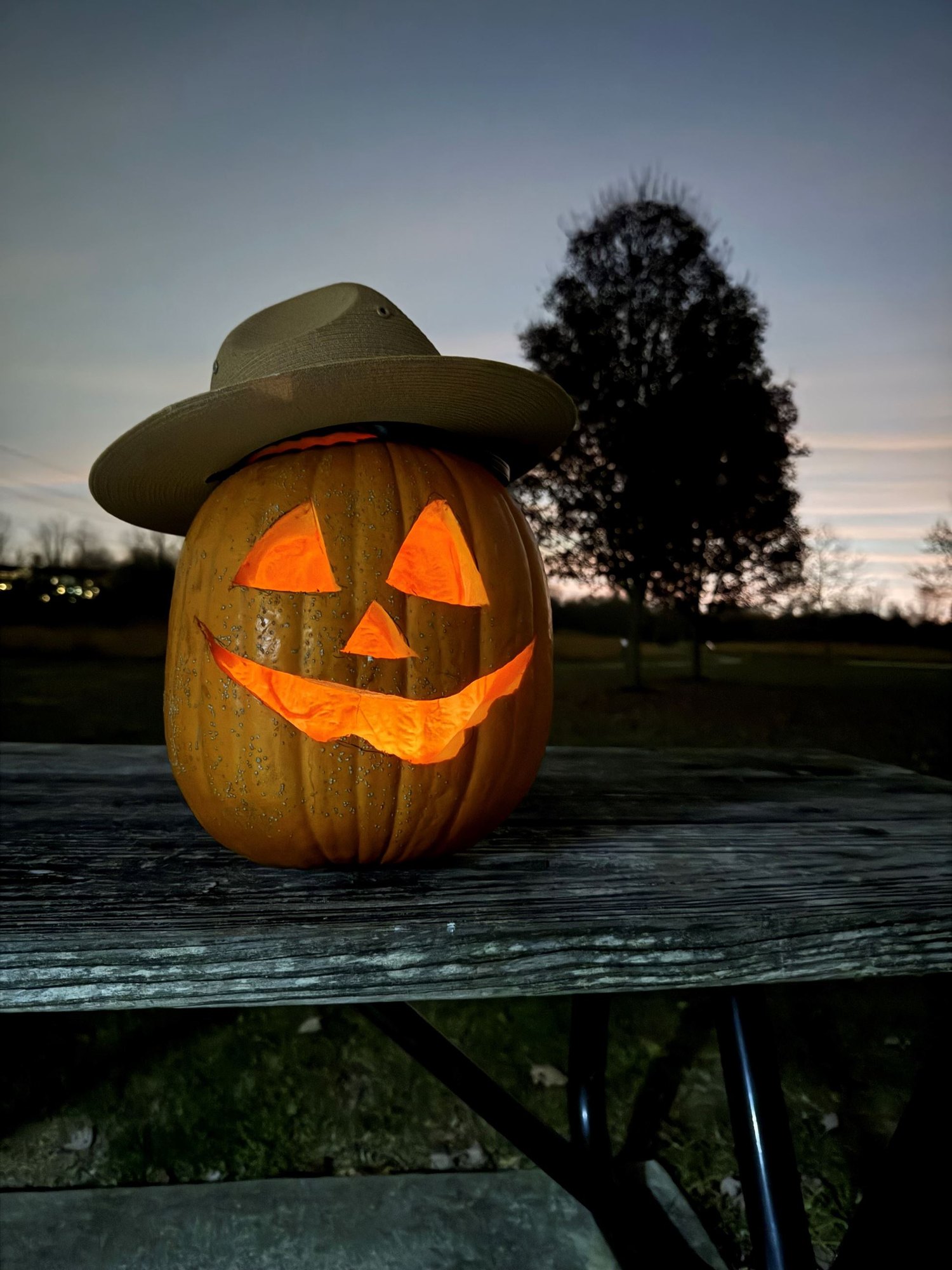 Lit carved pumpkin wearing a wide-brimmed ranger hat on an outdoor picnic table at dusk.
